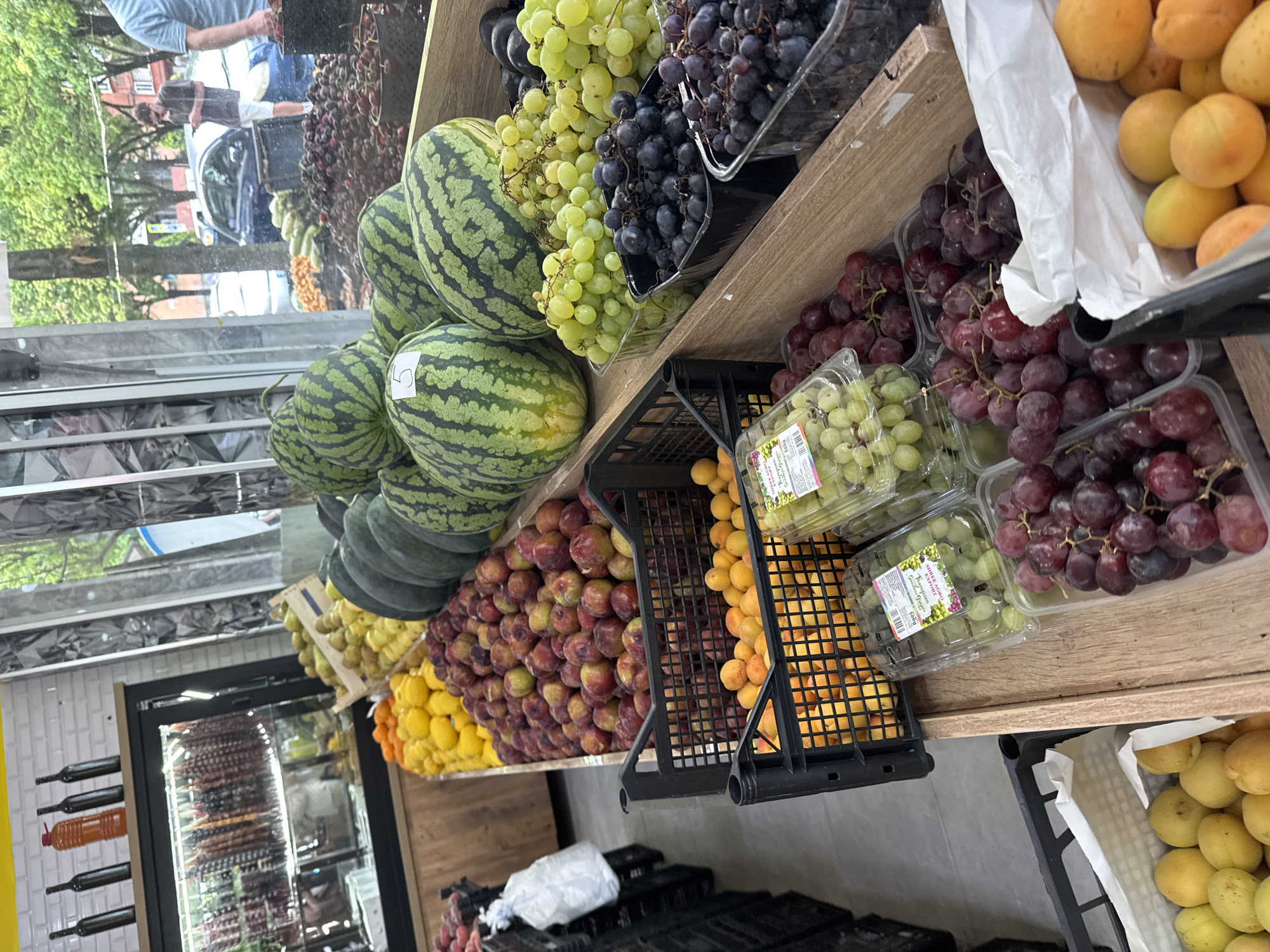 This is a close-up, vertical shot of a fruit stand in Batumi, Georgia, featuring a variety of fresh produce. In the foreground, several plastic containers and black crates are filled with grapes, both green and dark purple, as well as red plums and bright orange apricots. Behind these, large watermelons are displayed, some with price stickers. Further back, more fruit is visible, including yellow lemons and pears. The stand appears to be indoors, with a glass display case showcasing cured meats in the background. Through a window or open doorway, a street scene with trees, cars, and pedestrians is visible, suggesting an urban setting. The lighting suggests it is daytime.
