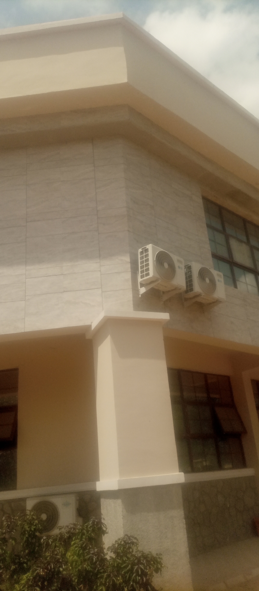 The image shows the exterior of a building with a modern design, featuring beige-colored walls and a lighter beige trim along the roofline. The building's facade is clad in light-colored, rectangular tiles with a subtle veining pattern, resembling marble. Two air conditioning units are mounted on the wall, one slightly higher than the other, both white with circular vents. Large, dark-framed windows are visible, some appearing to be open. A light beige pillar extends upwards from the ground level. In the lower portion of the image, a portion of an air conditioning unit is visible near some lush green foliage, suggesting a ground-level installation or a lower section of the building. The sky above is partly cloudy. The overall scene depicts a well-maintained building.