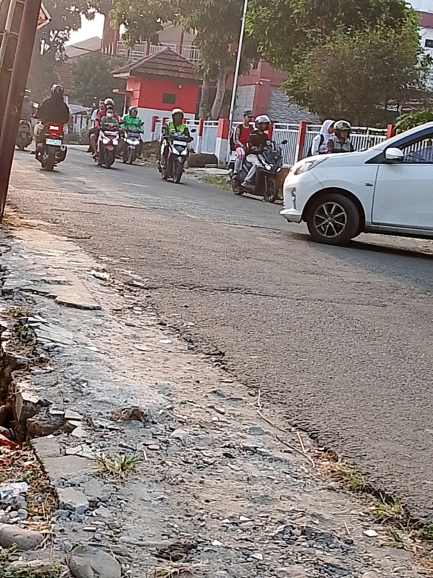 The image depicts a street scene in Cibinong, Indonesia, with a group of motorcyclists traveling along a paved road. The road appears to be in moderate condition, with some cracks and uneven surfaces visible along the side. On the left side of the road, there is a rough embankment with debris and some sparse vegetation.
In the foreground, a white car is partially visible, suggesting traffic on the road. Further down the road, several individuals are riding motorcycles. Some riders are wearing helmets and casual clothing, while others appear to be students based on their attire, including a person in a white hijab. The motorcyclists are moving in the same direction.
Behind the road, buildings with red roofs and white fences are visible, indicating a residential or commercial area. Trees with green foliage are also present, contributing to the overall urban landscape. The lighting suggests it might be early morning or late afternoon, with soft sunlight casting shadows. The presence of multiple people on motorcycles is a common sight in urban areas of Indonesia, reflecting local transportation patterns.