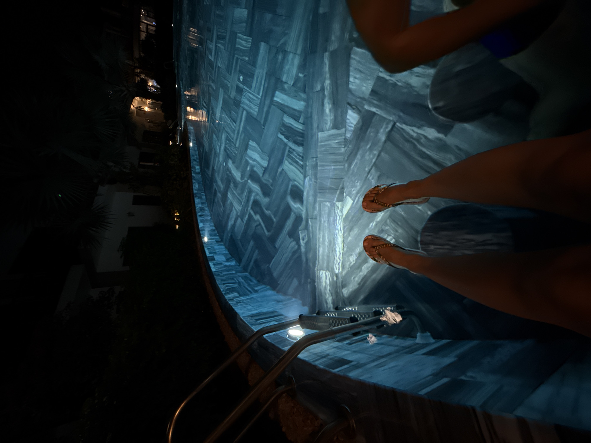 The image captures a nighttime scene at a swimming pool, likely in a resort setting in Phú Quốc, Vietnam. The pool's interior is illuminated with a soft blue light, revealing distinctive blue and grey patterned tiles on its floor.
In the foreground on the right, the tanned legs and feet of a person, wearing golden flat sandals, are positioned at the water's edge or slightly submerged. Further into the pool on the upper right, another individual's upper body and arms are visible, suggesting they are in the water or seated by the edge, wearing a blue swimsuit top. They appear to be relaxing or entering/exiting the pool.
On the left side of the pool, a metal ladder or set of steps provides access to the water, with small underwater lights visible. The surroundings are dark, with the faint silhouettes of buildings and tropical foliage, including palm-like leaves, visible against the night sky. Some faint lights from the buildings can be seen in the distance. The water appears calm and reflective. The overall atmosphere conveys a sense of relaxation and leisure during an evening swim or poolside moment. There is no visible text in the image.