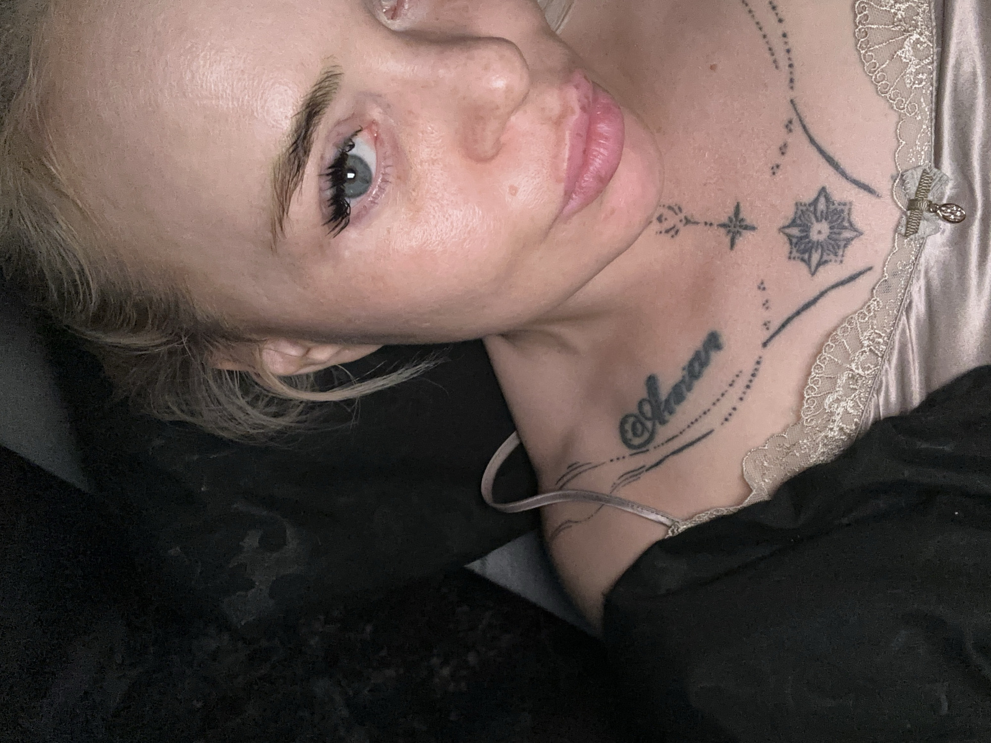 A close-up, high-angle portrait features a fair-skinned individual with blonde hair, visible from the forehead to the upper chest, captured in the city of Unknown, Unknown. The subject has light blue eyes with prominent eyelashes, a direct gaze, and full, pinkish lips slightly pursed. The forehead shows some sheen, and the cheeks exhibit minor skin variations. A light-colored, possibly champagne or pale pink, satin camisole with a lace trim along the neckline is worn, featuring a thin spaghetti strap on the left shoulder. A small decorative bow with a dangling charm is affixed to the center of the lace trim. Tattoos are visible on the upper chest: script reading "Shaman" (or similar) on the right collarbone, and a vertical arrangement of geometric and floral symbols (including a star and a lotus-like design) on the sternum, framed by curved and dotted lines. A dark, possibly black, garment drapes over the left shoulder. The background is uniformly dark and appears to be an interior space with soft, frontal lighting.