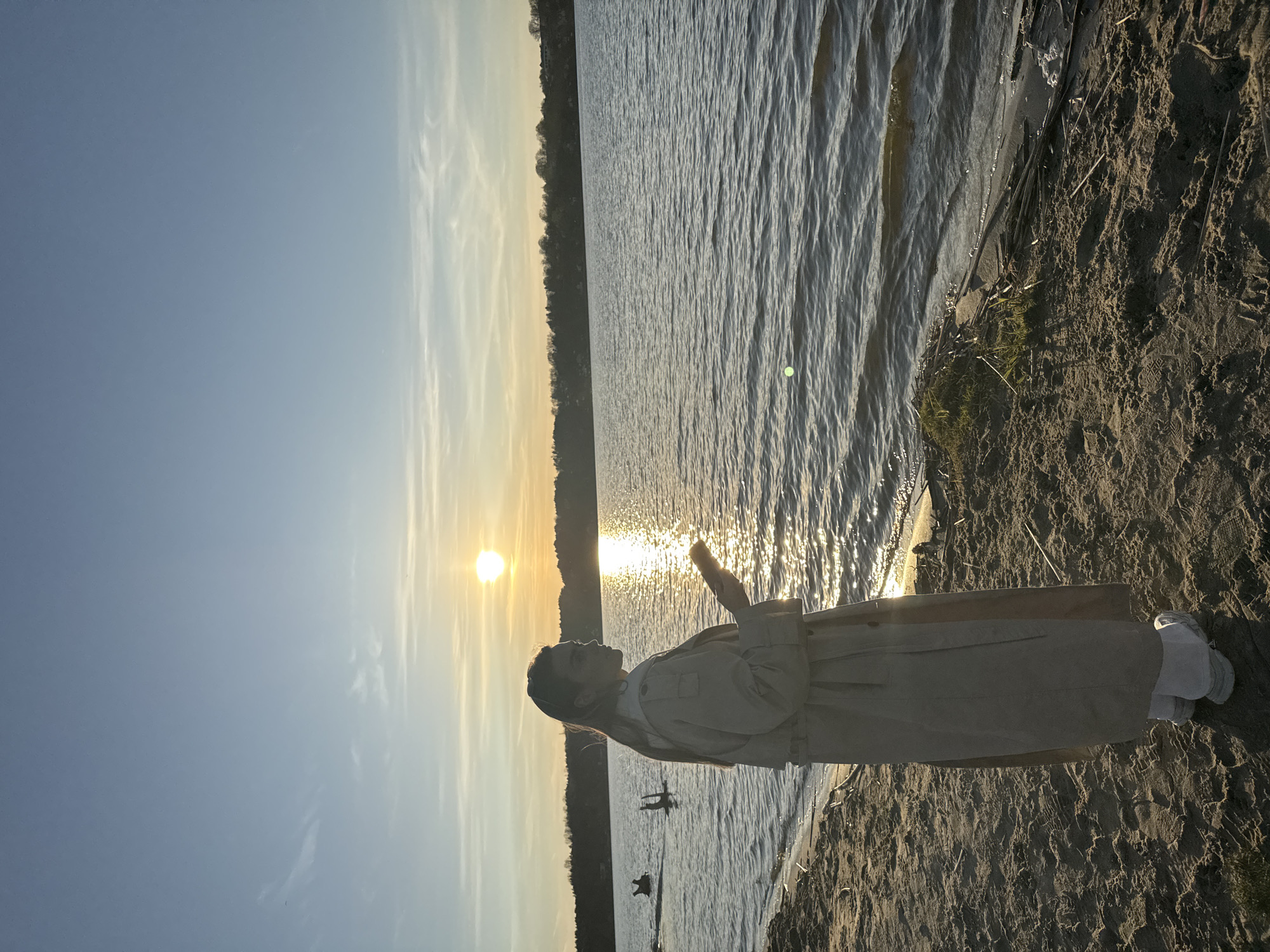 The image captures a serene sunset scene by a large body of water, likely a lake or reservoir, in Arciechów, Poland. The main subject is a person standing in profile on a sandy shore, looking directly towards the setting sun. They are dressed in a light-colored, possibly beige or off-white, trench coat or long jacket, with lighter trousers or shoes.
The sun is a prominent, bright golden orb positioned low on the horizon, slightly to the left of the frame's center. Its intense light casts a brilliant, shimmering path of reflections across the water, which shows gentle ripples. A dark treeline is visible in the distance, silhouetted against the bright sky, marking the opposite bank of the water body.
The sky displays a clear gradient from a soft blue at the top to warm yellows and oranges near the sun, with a few wispy clouds streaking across. The foreground features a sandy beach with visible textures and some scattered debris or vegetation near the waterline. The lighting suggests the "golden hour," creating long shadows and a warm, tranquil atmosphere. The person appears to be enjoying or contemplating the peaceful moment. No visible text is present in the image.