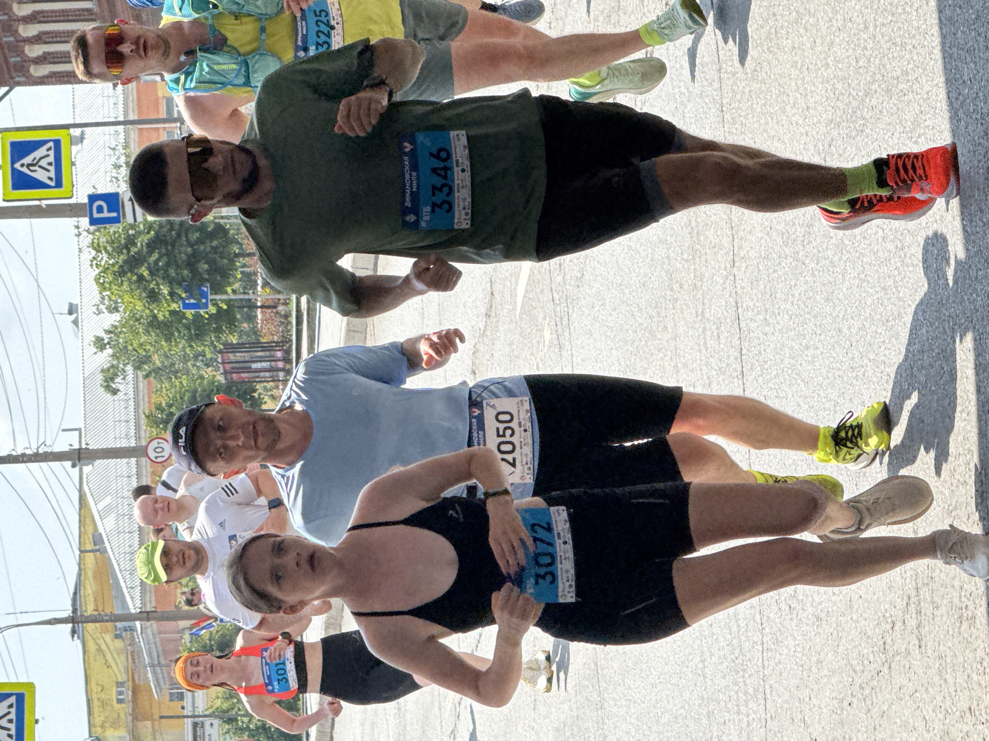 The image depicts a daytime outdoor running race in Izhevsk, Russia. In the foreground, three runners are visible in motion. The woman on the left, wearing a black sports bra and black shorts, has race number 3072 pinned to her front. To her right, a male runner in a light blue t-shirt and black shorts has race number 2050. Further to the right, another male runner in a dark green t-shirt and black shorts, wearing sunglasses, displays race number 3346. Behind them, other runners and spectators are present. The background reveals urban elements, including buildings, trees, traffic signs, and utility poles, under a bright sky. The race appears to be on an asphalt road surface.