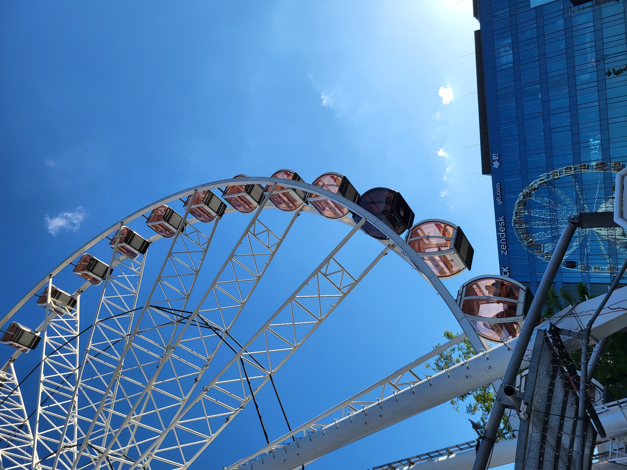 A large observation Ferris wheel dominates the left and upper-left portion of the image, set against a clear, deep blue sky with a few scattered white cumulus clouds. The wheel's structure is white, composed of numerous triangular metal trusses forming its frame and spokes. Approximately 12 to 15 enclosed passenger cabins are visible, primarily reddish-amber or tinted brown, hanging from the wheel's circumference. One cabin in the lower-left-mid section contains at least two indistinct human silhouettes.
In the mid-ground and background, filling the right side of the frame, stands a modern, multi-story building with a reflective, blue-tinted glass facade. Several company logos and text are visible on the building: "CK zendesk" is clearly identifiable on the upper left portion of the facade, followed by "gft.com" to its right, and "DP2" further to the right. The building's glass facade reflects a partial image of the Ferris wheel and the blue sky. The top of the building features a flat roofline with several small antennae or protrusions.
Bright sunlight creates a lens flare or sun glow in the upper right corner of the sky. In the immediate lower-left foreground, partially obscured, are grey metal support structures, including horizontal bars, mesh, and platform elements associated with the base of the Ferris wheel. Some green foliage from a tree is visible through the lower spokes of the wheel.
The scene is located in **Wrocław, Poland**.