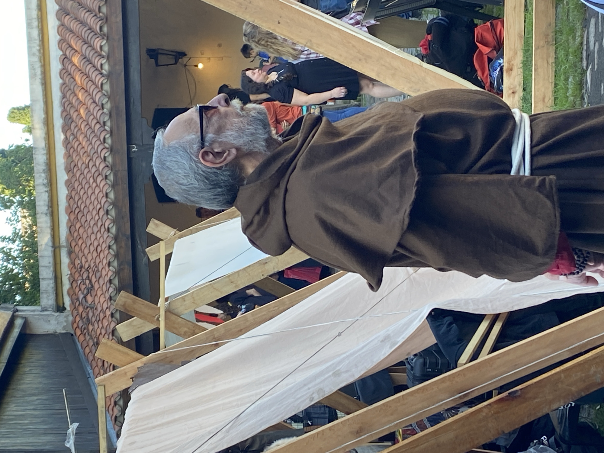 A medium, long shot depicts a man wearing a brown robe, possibly a religious figure, standing outdoors in daylight. The man is gray-haired and wearing glasses. He is standing amidst a wooden structure covered with white fabric. Other people are in the background, some sitting. The surrounding environment appears to be urban, with a building featuring terracotta tile roofing visible. While not explicitly stated, the query indicates that the location is in Buenos Aires, Argentina. The exact activity and relationship between the man in the robe and the surroundings are not immediately clear.