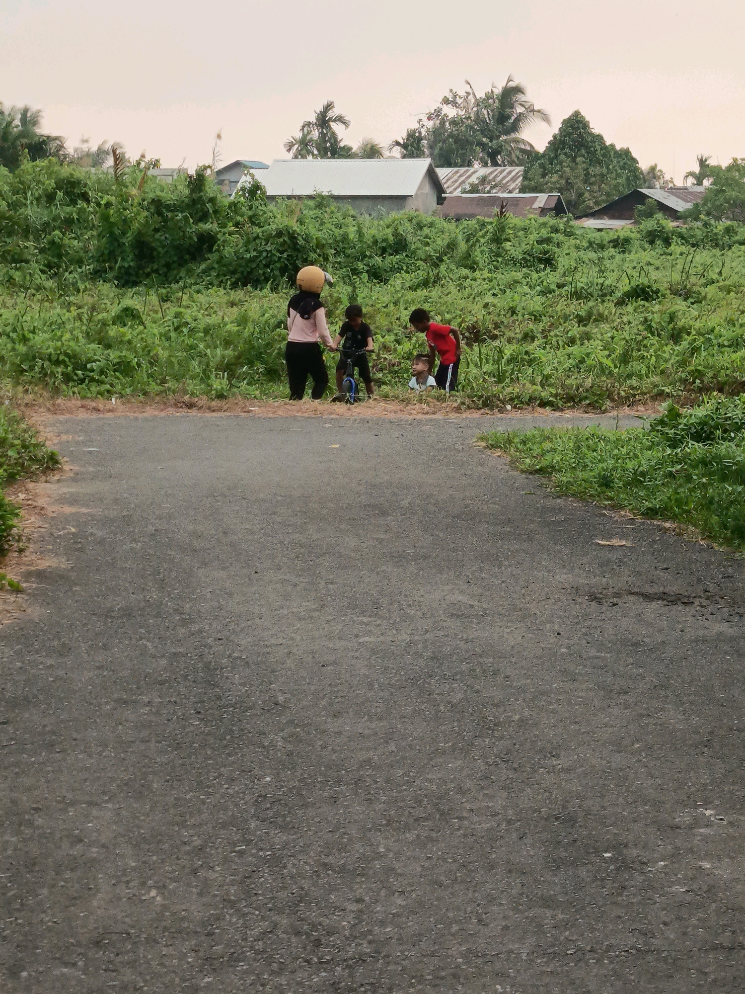 The image shows a group of children standing by a paved road in a rural area of Banjarmasin, Indonesia. The children appear to be playing or interacting near a patch of tall grass. In the background, there are several houses with corrugated metal roofs, surrounded by lush vegetation and trees, including palm trees. The sky is overcast, suggesting it might be early morning or late afternoon. The scene depicts a common everyday occurrence in the outskirts of the city.