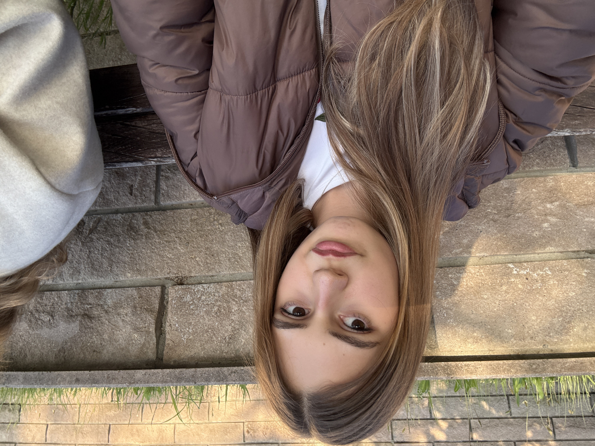 A young woman with long brown hair and brown eyes is sitting outside in an urban environment. She is wearing a brown puffer jacket over a white shirt. She is sitting on a stone bench that is adjacent to a brick wall. Some vegetation is visible growing between the bricks. This location is possibly within the city of Panagyurishte, Bulgaria.