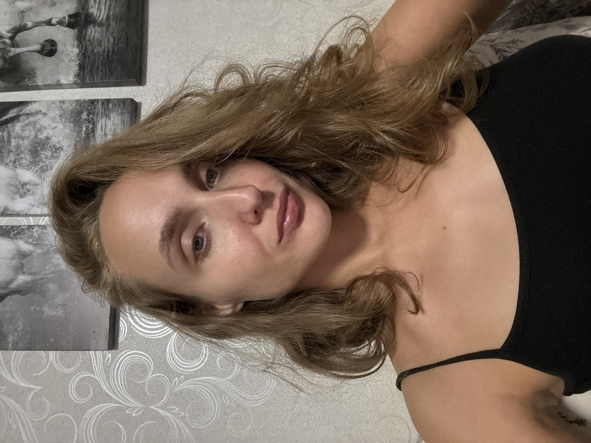 A woman with long, wavy blonde hair is looking directly at the camera, her head tilted slightly to her left. She is wearing a black tank top. Her skin is fair, with a faint blush on her cheeks. She has blue eyes and light pink lips. Behind her, the wall is covered with a light gray wallpaper featuring a swirling pattern. Above her, three framed pictures are hanging on the wall. The leftmost picture is partially visible, showing a horse in motion. The middle picture clearly depicts a horse splashing through water, and the rightmost picture also shows a horse in water, viewed from the back. The lighting in the image is soft, suggesting an indoor setting. There is no indication that this image was taken in Kuyeda, Russia, or any specific city.