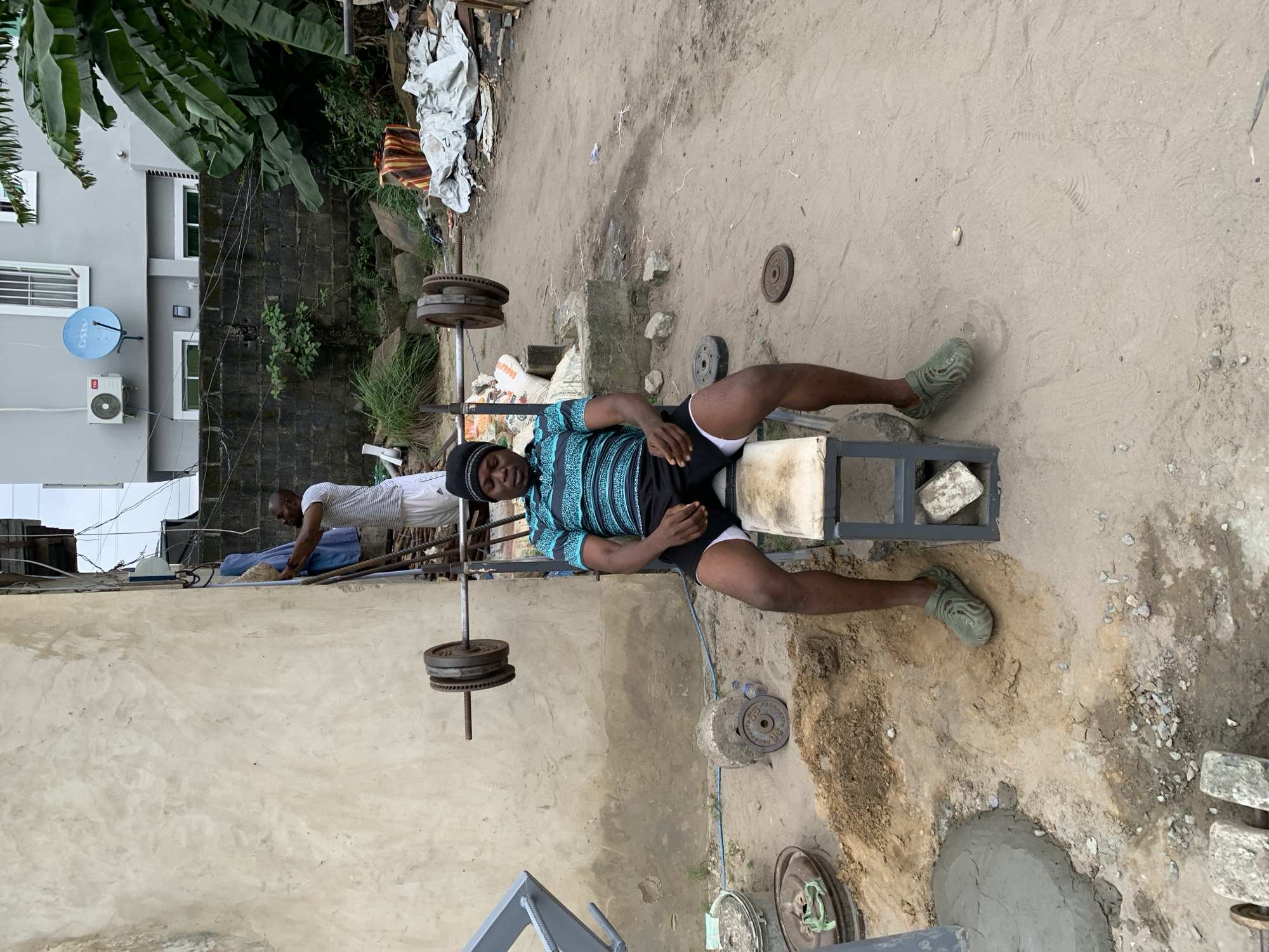 In Ado, Nigeria, a man is seated on a makeshift weight bench, focused on his workout. He wears a black beanie, a teal and black striped t-shirt, and black shorts with white trim. A barbell with weights rests on a rack behind him. To his right, another man, wearing a white t-shirt and white shorts, is hanging a blue cloth on a rack attached to a rough-textured wall. The environment is an outdoor setting with a dirt ground, scattered debris, and a partially constructed building in the background. The scene appears to be a residential or informal outdoor gym area.