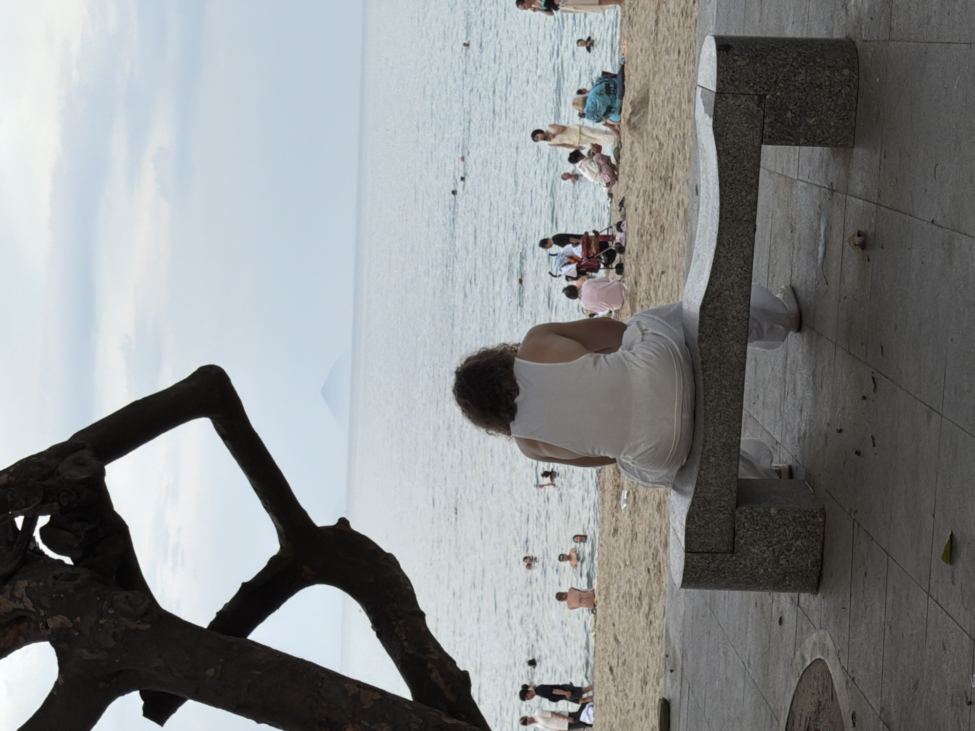 In Nha Trang, Vietnam, a person is seen from behind sitting on a stone bench facing the ocean. The individual wears a white tank top and white pants, with curly dark hair. Behind them, the sandy beach is populated by several people, some swimming in the ocean, others relaxing on the sand. A tree branch extends into the frame from the left, partially obscuring the view of the distant horizon where a mountain peak is visible. The sky is overcast. The scene is captured from a low angle, emphasizing the bench and the seated figure.