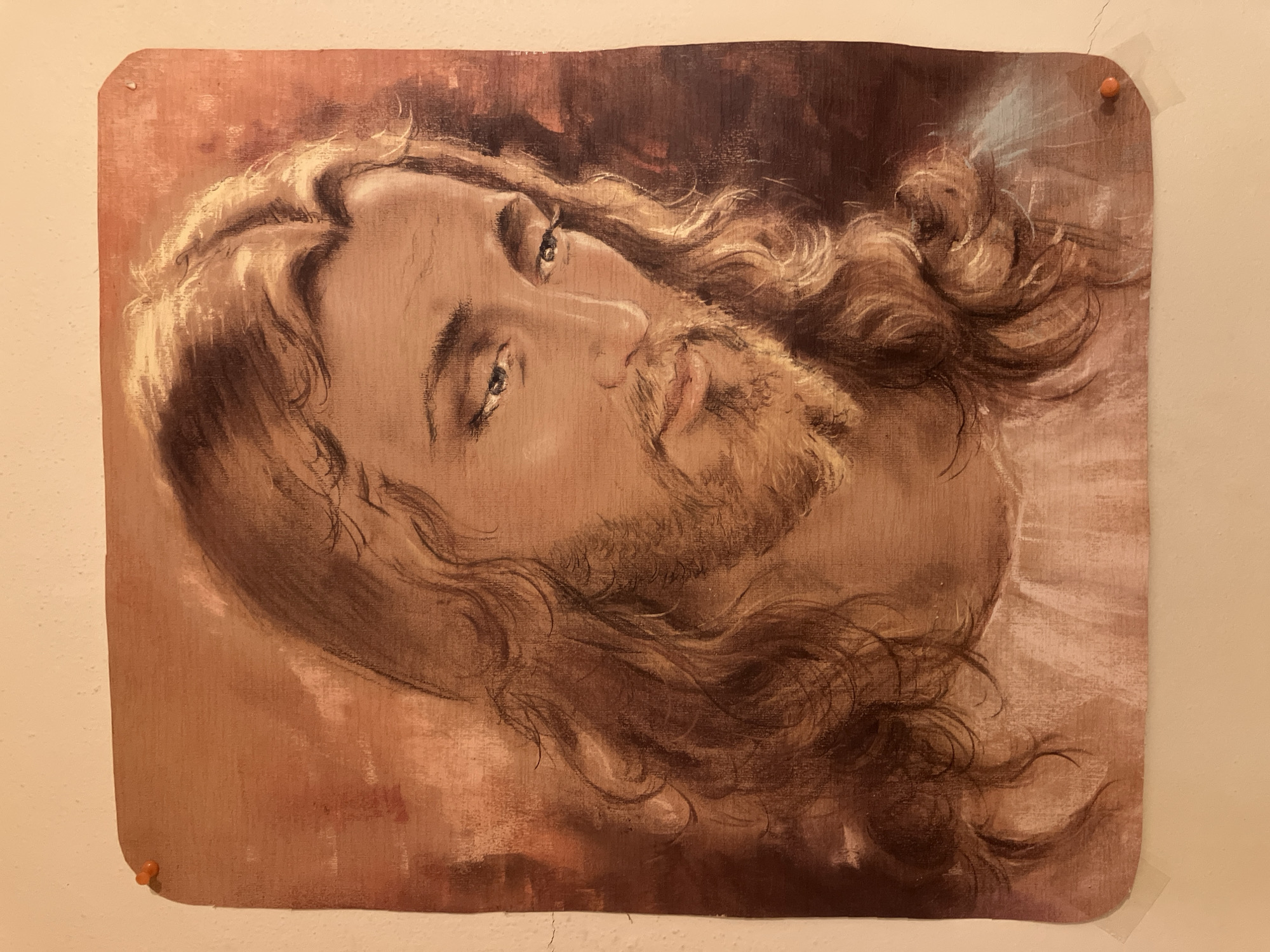 The media file presents a portrait-style artwork depicting the head and shoulders of a male figure, strongly resembling traditional depictions of Jesus Christ. The figure has long, wavy light brown hair and a beard, with a serene and contemplative expression. His eyes are cast slightly upwards and to the viewer's left.
The artwork is rendered in warm, muted tones, primarily browns, beiges, and subtle reddish-orange hues. The background within the artwork is abstract, featuring swirling brushstrokes, with a hint of ethereal light, possibly blue or white, emanating from behind the figure's head on the right side. The figure's head is tilted within the frame, appearing rotated slightly clockwise.
The artwork itself is a physical print or painting, mounted on a plain, light-colored wall, likely off-white or beige. It is secured to the wall by two orange pushpins, one visible in the top right corner and another in the bottom left. A small piece of clear tape is also visible adhering the bottom right corner of the artwork to the wall. The corners of the artwork are subtly rounded. Faint hairline cracks are noticeable on the wall surface, particularly near the top right pushpin and along the bottom edge of the image. There is no visible text on the artwork or the wall.