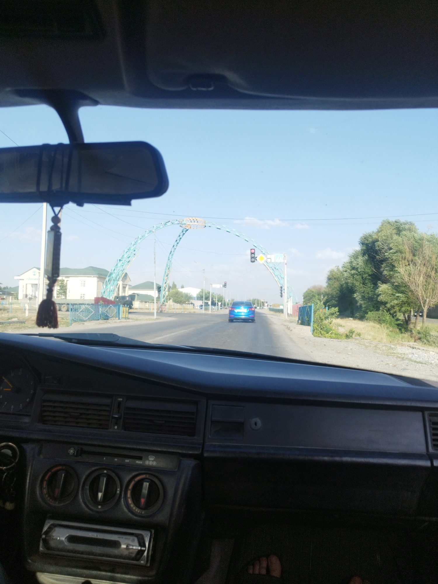 The image is taken from inside a car looking forward through the windshield. The view shows a street scene in the village of Koksayaq Rural District, Kazakhstan. In the foreground, the car's dashboard and rearview mirror are visible. A car air freshener hangs from the rearview mirror. Ahead, a blue car is driving on the road. A large, turquoise archway spans the road, supporting traffic lights and pedestrian crossing signs. On the left side of the road, there are buildings and a fence. On the right side, there are trees and dry vegetation. The sky is clear and blue.