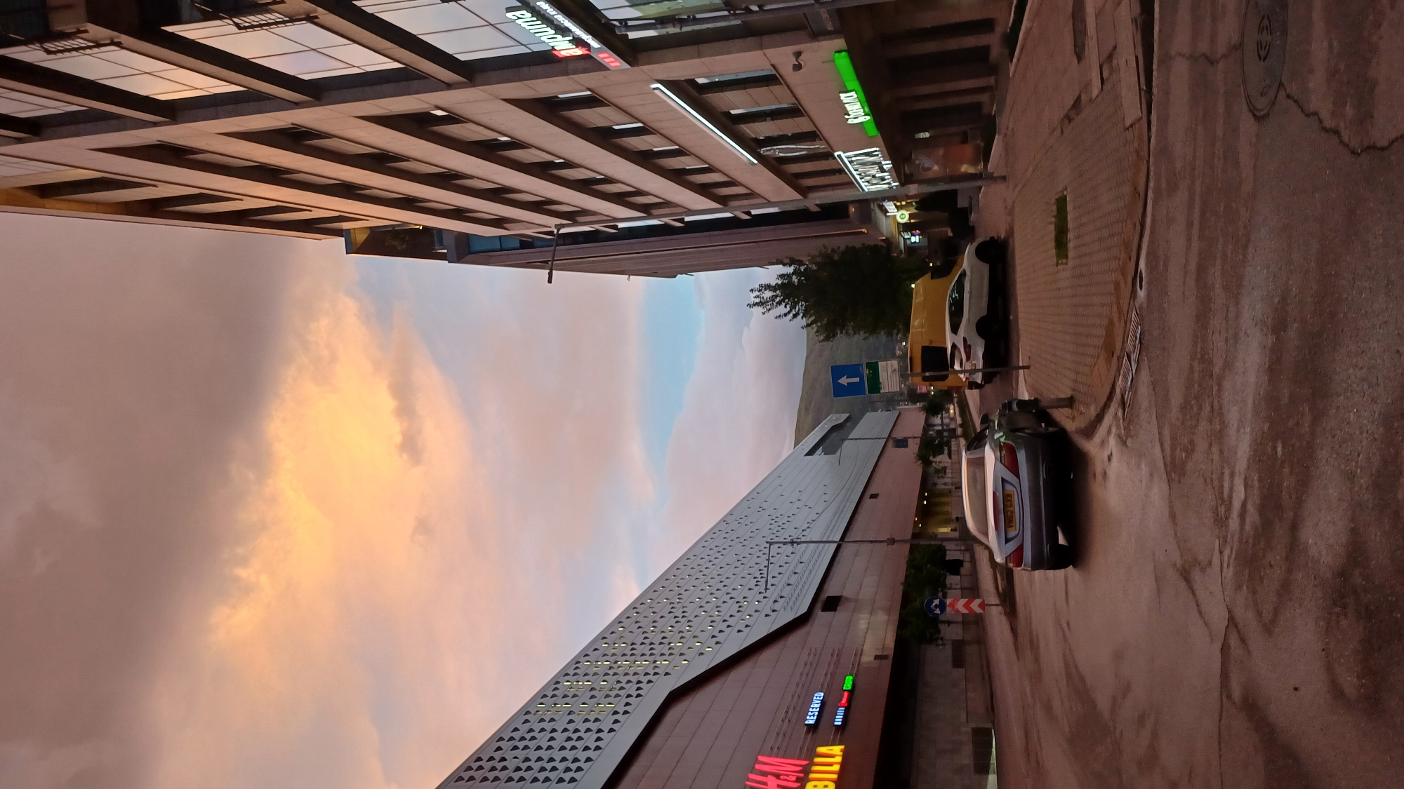 The image depicts an urban scene in Sofia, Bulgaria at dusk. The sky is a mix of blues and pinks/oranges from the setting sun. Two large buildings dominate the scene: one a modern, angled structure with multiple small, triangular windows, bearing the signs "H&M" and "BILLA", and the other a taller, multi-story building with a repetitive window pattern and a commercial sign "eunda" on top.
A street runs between the buildings, with two cars parked along the right side. One is a grey sedan and the other is a white and yellow van. A street sign is visible near the van, directing traffic to the left. The street surface is dark and appears slightly wet, suggesting a recent rain or cleaning. A manhole is visible in the upper right corner.