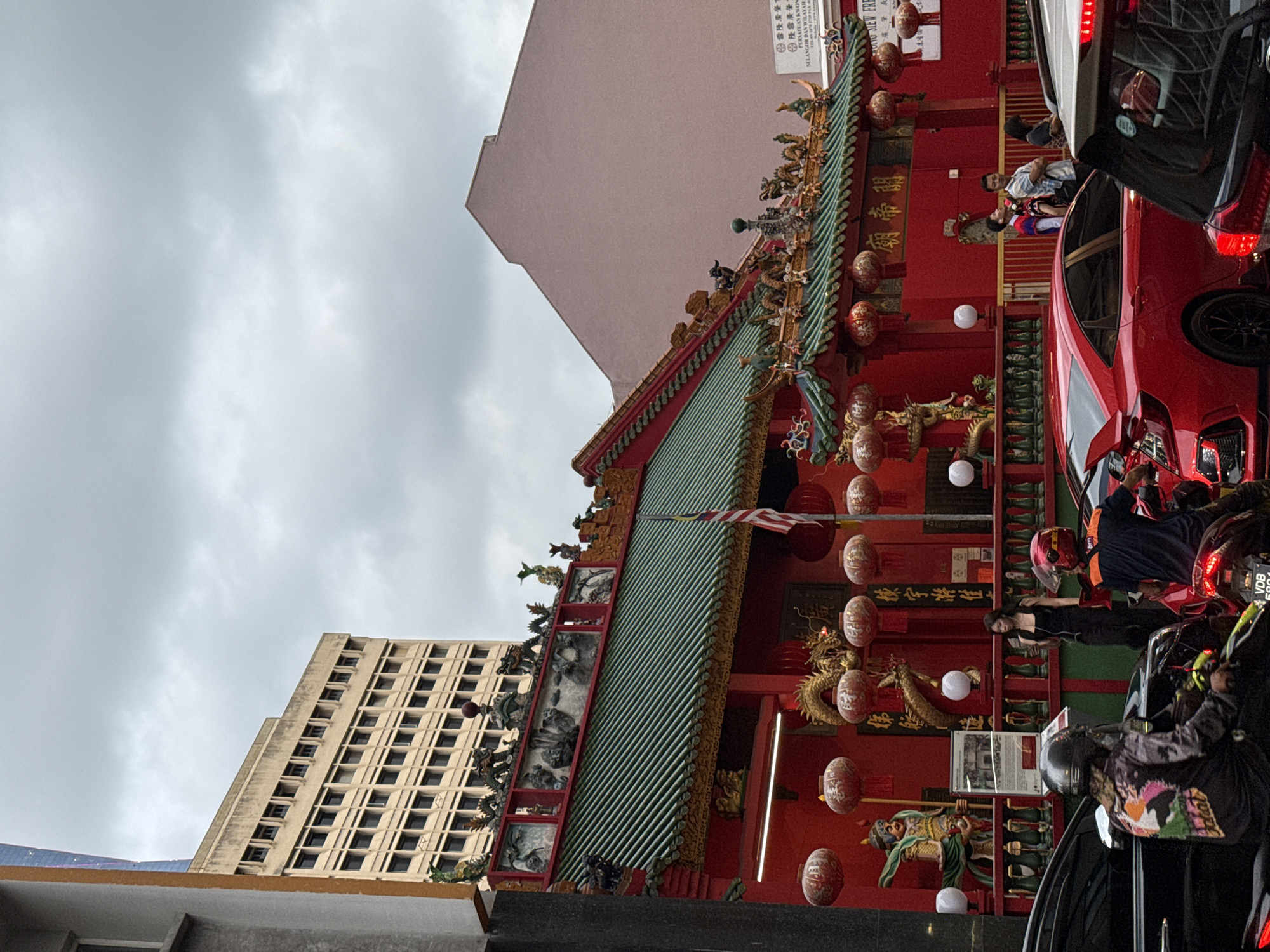 Here's a concise, matter-of-fact description of the image:
The image shows the Sin Sze Si Ya Temple in Kuala Lumpur, Malaysia, partially obscured by cars and people on the street. The temple is a traditional Chinese building, painted red, with a green tiled roof decorated with figurines. Red lanterns hang along the front of the temple. A Malaysian flag is visible hanging under the roof. A tall, modern building is seen in the background to the left, and a plain pink building is above the temple. People and vehicles are on the street in front of the temple. Text in Chinese and Malay can be seen on the temple walls.