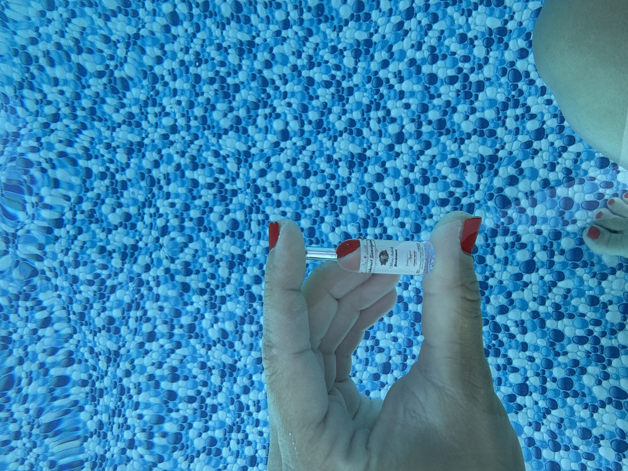 An underwater shot of a person's hand holding a small glass bottle with a red-capped stopper is captured against a blue tiled swimming pool background. The bottle has a label with text that is partially legible, appearing to indicate "Cabernet Sauvignon" and "Bordeaux". The person's fingernails are painted red. The background features a repetitive pattern of blue and lighter blue circular and oval shapes, characteristic of a swimming pool's tiled bottom. The image does not provide any information about the city of Gara Oreshets, Bulgaria.