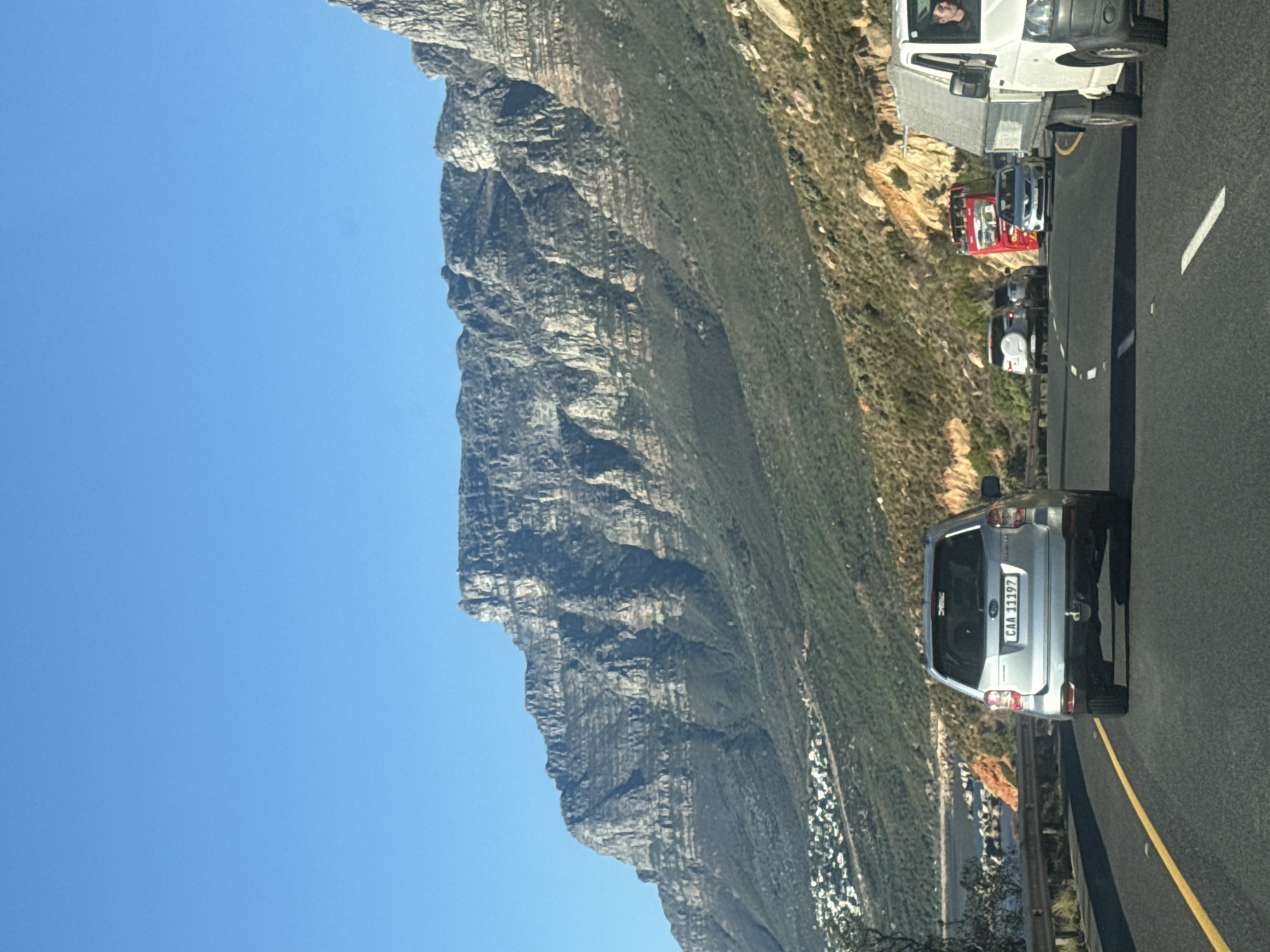 The image captures a scenic drive along a mountain road under a clear blue sky. In the foreground on the right, the rear of a silver SUV, bearing the South African license plate "CAA 11197" and a small "DRMC" sticker, is visible as it travels away from the viewer. Behind it, a red vehicle and a white delivery van follow on the two-lane asphalt road, which is marked with a solid yellow line on the left and a broken white line separating the lanes. A person's face is discernible in the driver's seat of the white van.
To the left, a massive, rugged mountain range dominates the scene, showcasing layered rock formations and patches of green vegetation clinging to its steep slopes. The sky above is a vibrant, cloudless blue, indicating bright, sunny weather and a daytime setting. On the right side, opposite the mountain, the landscape slopes downwards, covered in green scrub, with a scattered cluster of white buildings visible in the distant valley, suggesting a town or settlement. The "CAA" prefix on the license plate confirms the location as Cape Town, South Africa, likely a coastal route alongside mountains such as Table Mountain or the Twelve Apostles.
