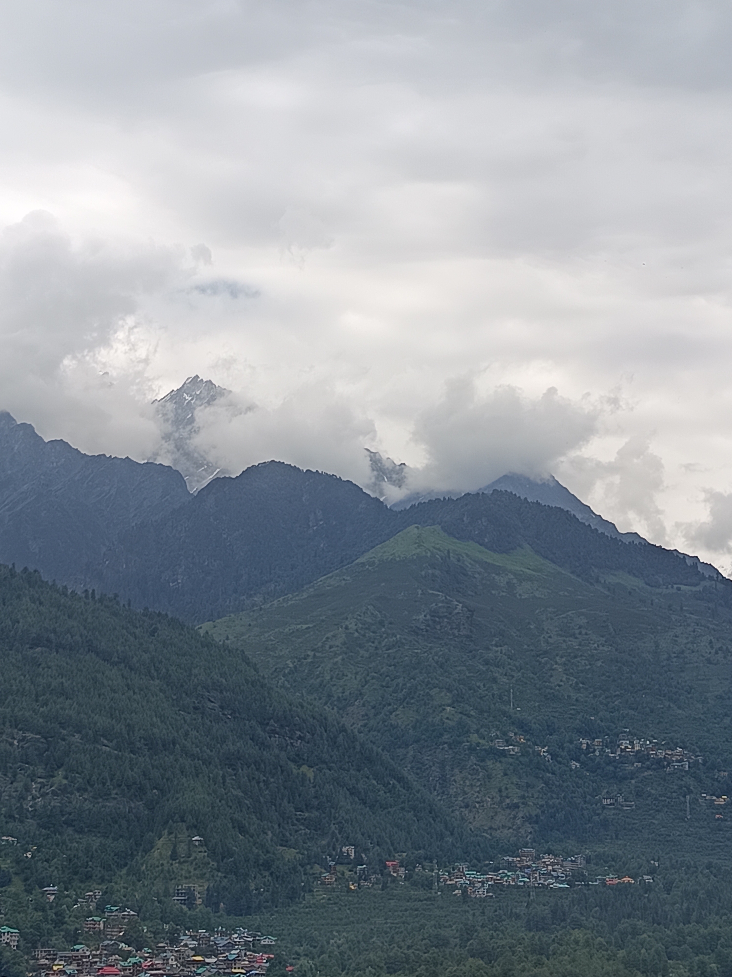 The image shows a mountain landscape with a village nestled in the foothills. The mountains are covered in lush green trees, with some snow-capped peaks visible in the distance. The sky is overcast with clouds. The village, identified as Vashisht, India, is characterized by clusters of buildings with colorful roofs, spread across the slopes. There are no people or specific actions depicted in the scene. The environment is natural and mountainous, with human settlement integrated into the landscape.