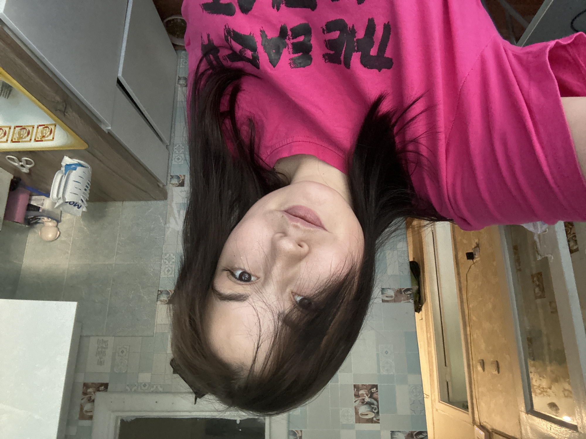 The media file is an indoor, eye-level, medium shot of a young woman with long, dark hair, wearing a bright pink t-shirt with black text. The text on the t-shirt appears to read "THE EAST". She is looking directly at the camera with a neutral expression.
The background suggests a domestic setting, likely a kitchen or bathroom, with tiled walls and what appear to be kitchen cabinets. On a counter to the left, there are some items including a white object with blue writing, possibly a cleaning product or food packaging, and a small pink bottle. The lighting is not clearly indicative of a specific time of day. The overall impression is a candid snapshot taken in a home environment. There are no clear indicators of weather or a specific event. The location is Chaikovsky, Russia.