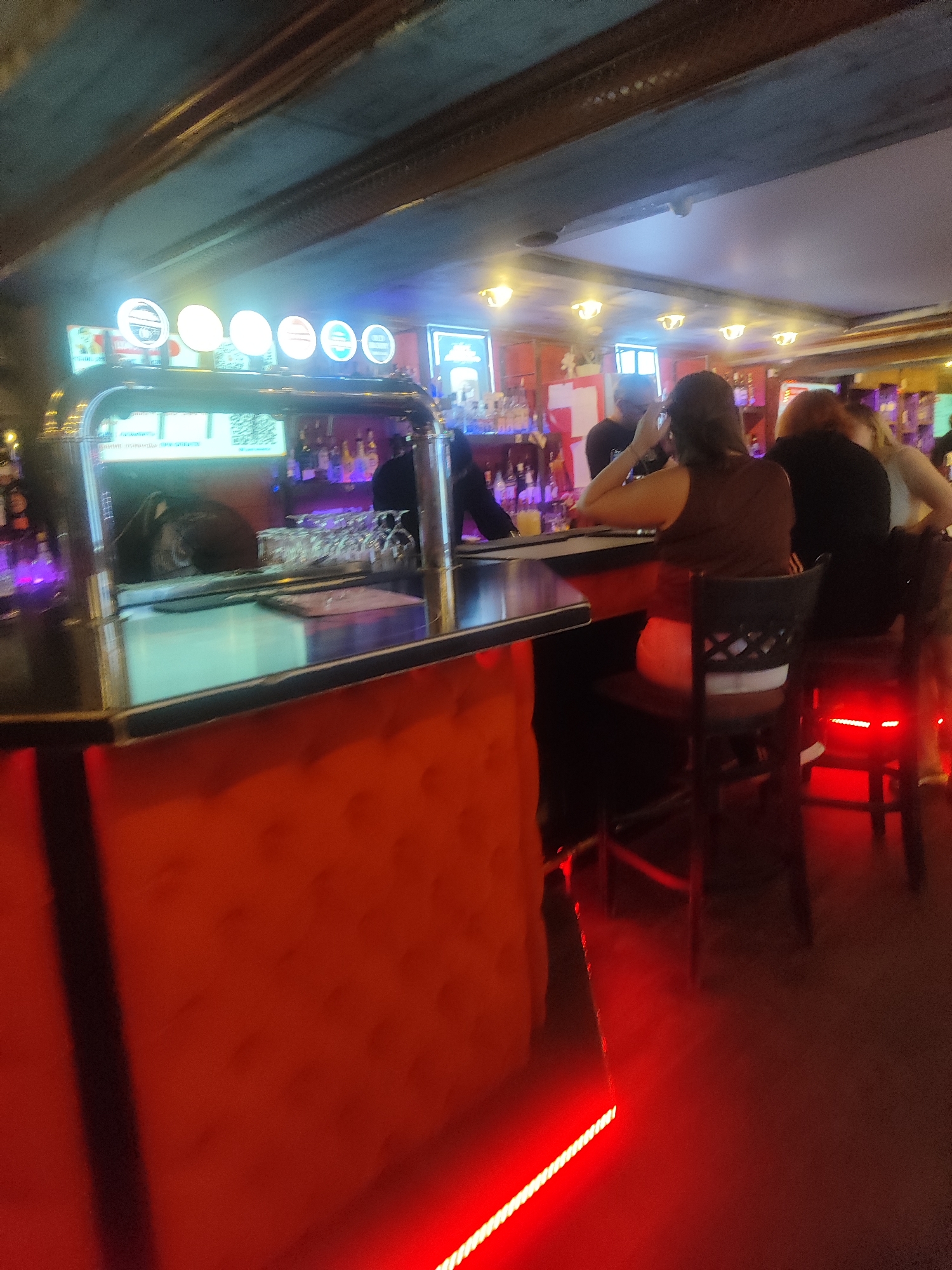 The image depicts the interior of a dimly lit bar, likely a pub or restaurant, in an unknown location. In the foreground, a portion of a textured orange bar front is visible, with red LED lighting strip along its base. A black countertop runs along the top of the bar, with a chrome beer tap system on the left side. Several people are seated at the bar, with their backs to the camera, appearing to be patrons. One person in the center is wearing a brown sleeveless top and a white and black striped skirt. To the right, another person is seen in a black top. The background shows a well-stocked bar with various bottles and glassware, illuminated by soft lighting. The ceiling is dark, with exposed beams and overhead lights. The overall atmosphere is casual and social, typical of a bar setting.