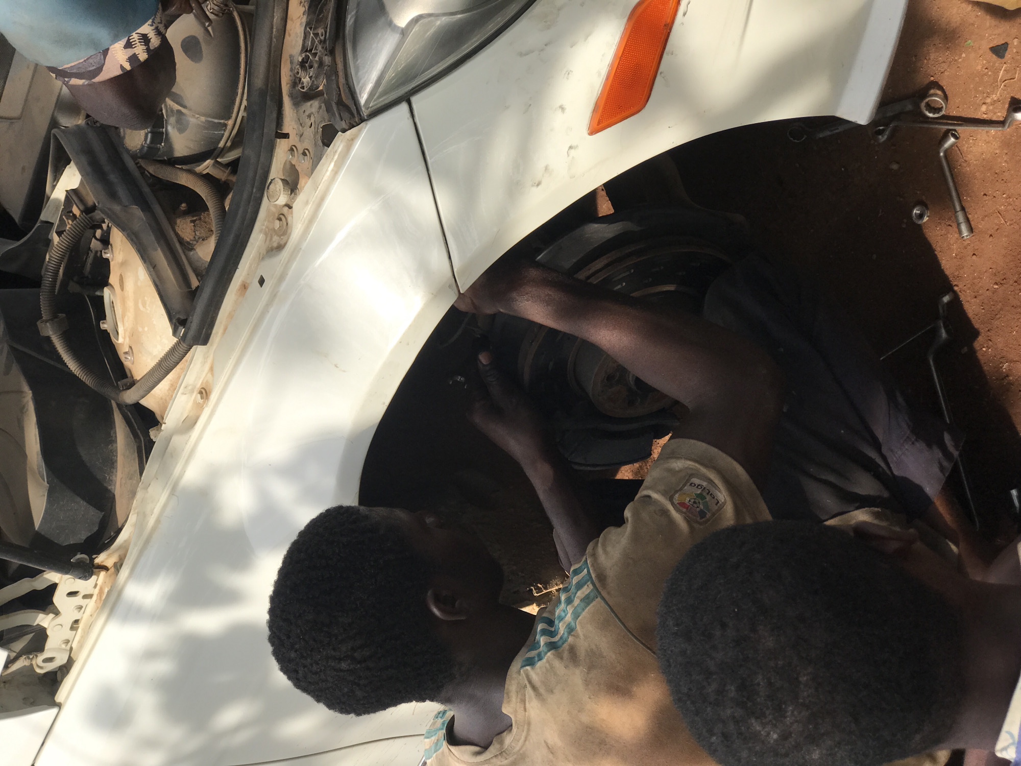The image captures an outdoor auto repair scene, likely in Abuja, Nigeria, during bright daylight. The primary focus is on the front-right side of a white car, possibly a sedan or SUV, where two individuals are actively engaged in mechanical work.
In the foreground, a person with dark skin and short, black hair is seen wearing a light-colored t-shirt with blue stripes. A "LaLiga" logo is visible on the right sleeve of their shirt. This individual is looking down into the car's wheel well, observing or assisting with the task. The dark-skinned arms of a second person are deeply inserted into the wheel well, suggesting they are working on components such as the suspension, brakes, or steering mechanism. A third person's head, partially obscured and wearing a patterned hat, can be seen at the top-left of the frame, near the car's engine bay.
The white car body shows signs of use and some dirt. The front-right headlight assembly and an orange side marker light are visible. The fender liner appears to be removed or pushed aside to allow access to the internal components. On the reddish-brown, dusty ground surrounding the car, various tools including wrenches, sockets, and loose nuts or bolts are scattered, indicating ongoing repair work. The bright, direct sunlight casts clear shadows, affirming it is daytime. The scene conveys a hands-on, focused effort to mend the vehicle in an informal outdoor garage setting.