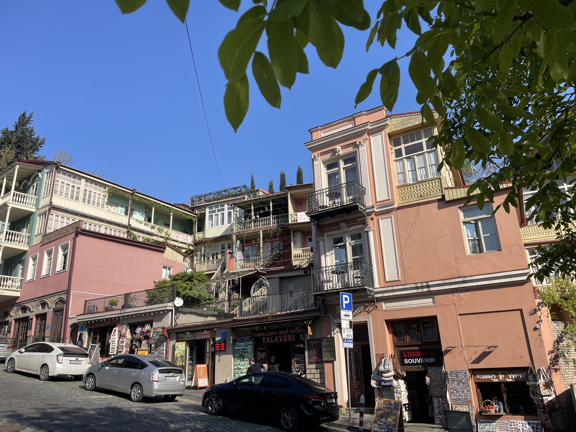 This media file captures a vibrant street scene in Tbilisi, Georgia, on a clear, sunny day. The view looks up towards colorful, multi-story buildings with traditional Georgian architecture, including numerous ornate balconies made of wood and wrought iron. Bright green leaves from an overhead tree partially frame the top of the image.
The buildings, painted in shades of pink, salmon, and light green, line a cobblestone street. At street level, several shops are visible. A prominent "TALAVERI WINE HOUSE" with Georgian script (ღვინის სახლი) and "SHOP BAR" occupies the center, displaying various items outside. To its right is a "SOUVENIR" shop, also with Georgian script (სუვენირი), featuring a wooden stand filled with postcards and other tourist items. An "EXCHANGE" shop is located to the left.
Three cars are parked on the street: two silver Toyota Priuses (license plates SU-797-RO and SG-771 SS) and a black sedan. A blue parking sign with a "P" symbol and a smaller blue sign marked "A 2166" are visible on the sidewalk. A few people can be faintly seen near the shops, engaging in daily activities. Balconies on the upper floors show signs of residential use, with some adorned with plants or clothes. The clear blue sky and strong shadows indicate it is likely late morning or early afternoon. A phone number, T: 557 17 48 53, is visible on a sign on one of the upper balconies, possibly indicating an apartment or property for sale.