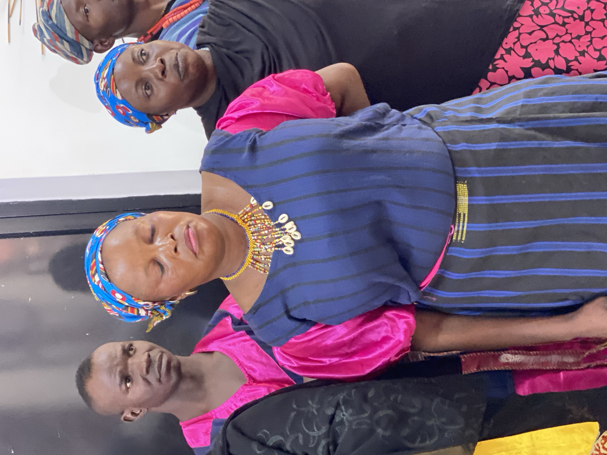 This image captures a group of people, likely women and one man, in what appears to be an indoor setting in Kwarau, Nigeria. The lighting suggests it could be daytime indoors or under artificial light.
In the foreground, a woman wears a blue and yellow patterned headscarf and a navy blue top with thin blue horizontal stripes. She also wears a prominent, multi-colored beaded necklace adorned with white cowrie shells around her neck. Her eyes are closed, and her lips are slightly parted, giving her a serene or contemplative expression. Beneath her blue top, a vibrant pink garment is visible, possibly part of her sleeves or an underlayer.
Behind her, to her left, another woman is visible. She wears a blue headscarf with red and yellow patterns, a black top, and an orange beaded necklace. She is looking directly at the camera with a neutral expression. Further back and to the top left, a third woman is partially seen, wearing a striped headscarf (blue, red, and white) and a black top, also looking towards the viewer.
On the bottom left, partially cropped, a man with a short haircut is visible, wearing a pink garment under a dark top, also looking towards the camera with a neutral expression. The subjects' attire, including patterned headscarves and beaded jewelry, suggests cultural or traditional clothing common in the region. The background consists of plain dark and light walls, providing a simple backdrop for the group. There is no visible text in the image.