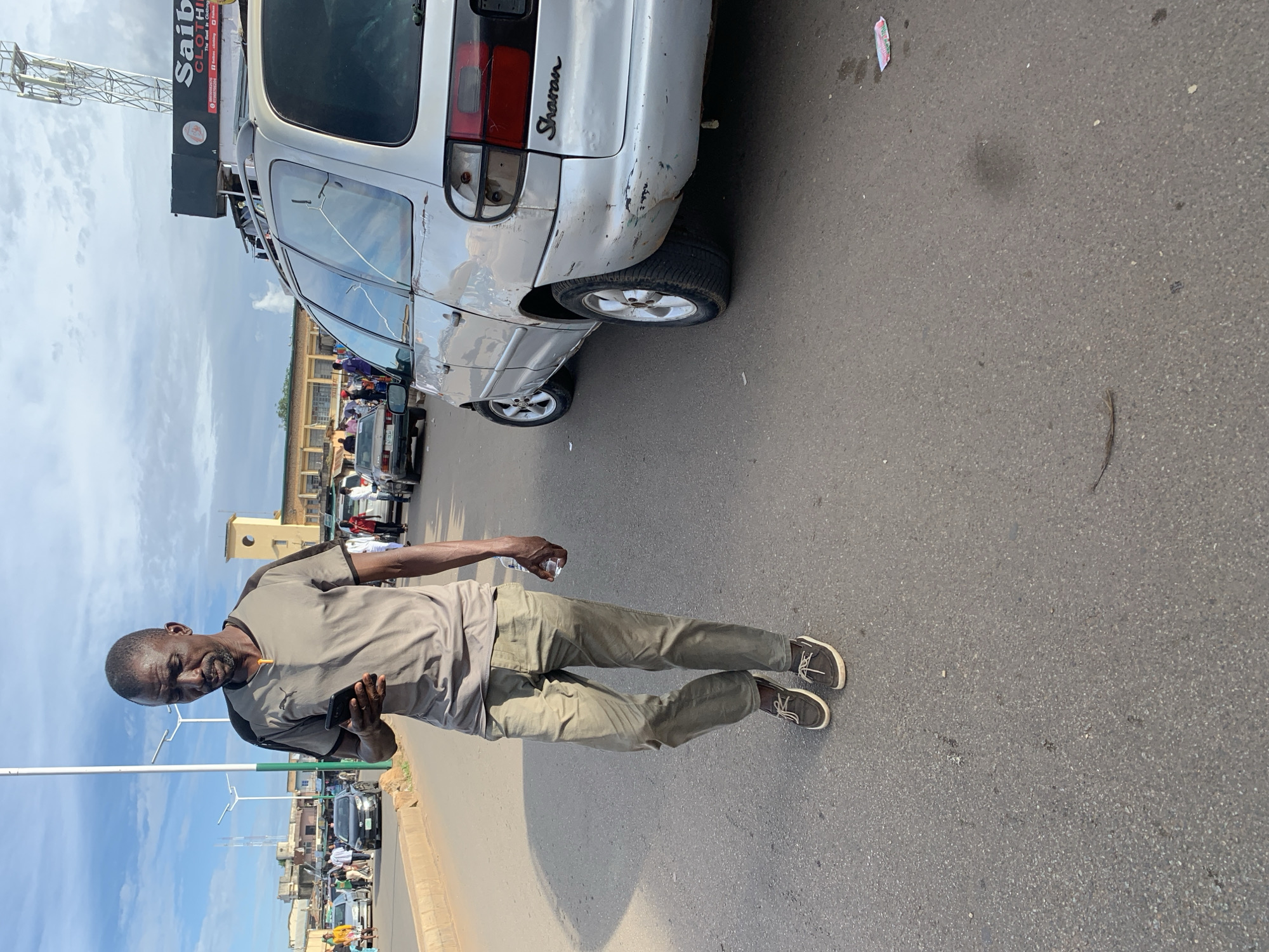 A man walks on a paved street in Jos, Nigeria, looking at his phone. He is wearing a grey t-shirt with black trim, light olive-colored pants, and brown boat shoes. He holds a clear plastic bottle in his right hand. To his right, a silver minivan is parked, showing signs of wear and tear, including dents and scuffs. The minivan has a black roof rack and a "Sharon" emblem on its rear. In the background, other vehicles and pedestrians are visible, as are buildings and a cell tower. The sky is partly cloudy. The scene appears to be an outdoor market or busy street.