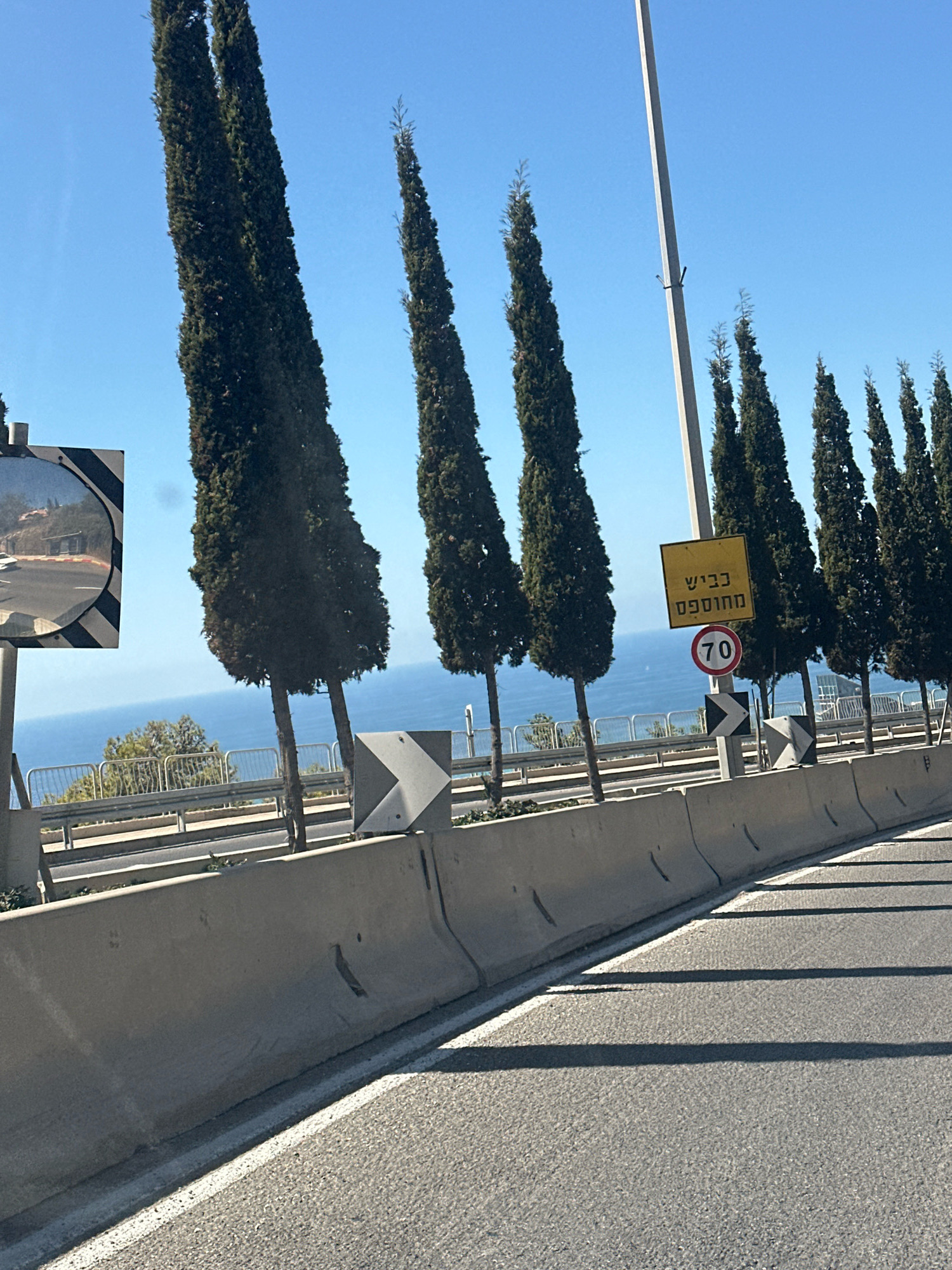 The image shows a view from a vehicle driving along a road in Haifa, Israel, with the Mediterranean Sea visible in the background. Tall, slender cypress trees line the right side of the road, casting long shadows on the asphalt. A concrete barrier separates the road from a lower level, beyond which is a metal fence and more vegetation.
Traffic signs are prominently displayed. A yellow square sign with black Hebrew text reading "כביש מחלף" (meaning "interchange road") is mounted on a pole. Below it is a circular red and white speed limit sign indicating "70". Further to the left, a large convex mirror is mounted on a pole, reflecting the road and surrounding area. A chevron sign, indicating a change in the road's direction, is visible to the left of the speed limit sign. The sky is clear and blue.