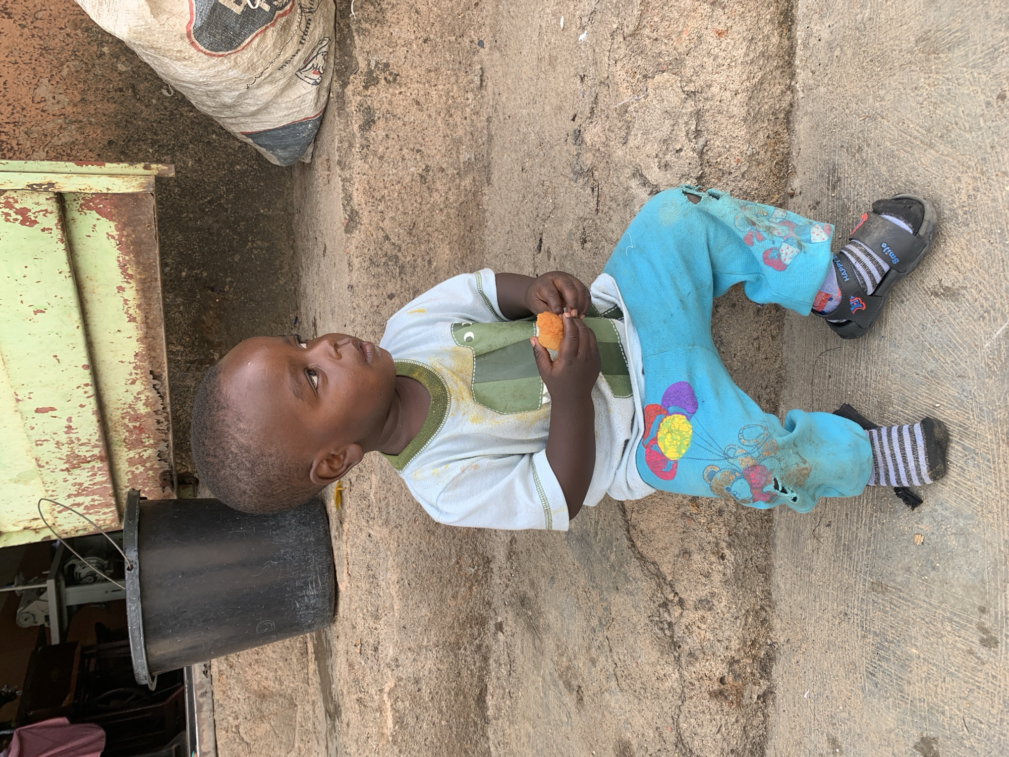 A young child is sitting on a concrete surface, holding a piece of food, potentially bread. The child is wearing a light-colored t-shirt, blue pants with colorful designs, striped socks, and black sandals. To the left of the child is a dark bucket and a green metal object, potentially a container or door. In the upper left corner is a large, possibly filled, sack. The location is implied to be in the city of Jos, Nigeria. The concrete surface is cracked and stained, suggesting an outdoor environment.