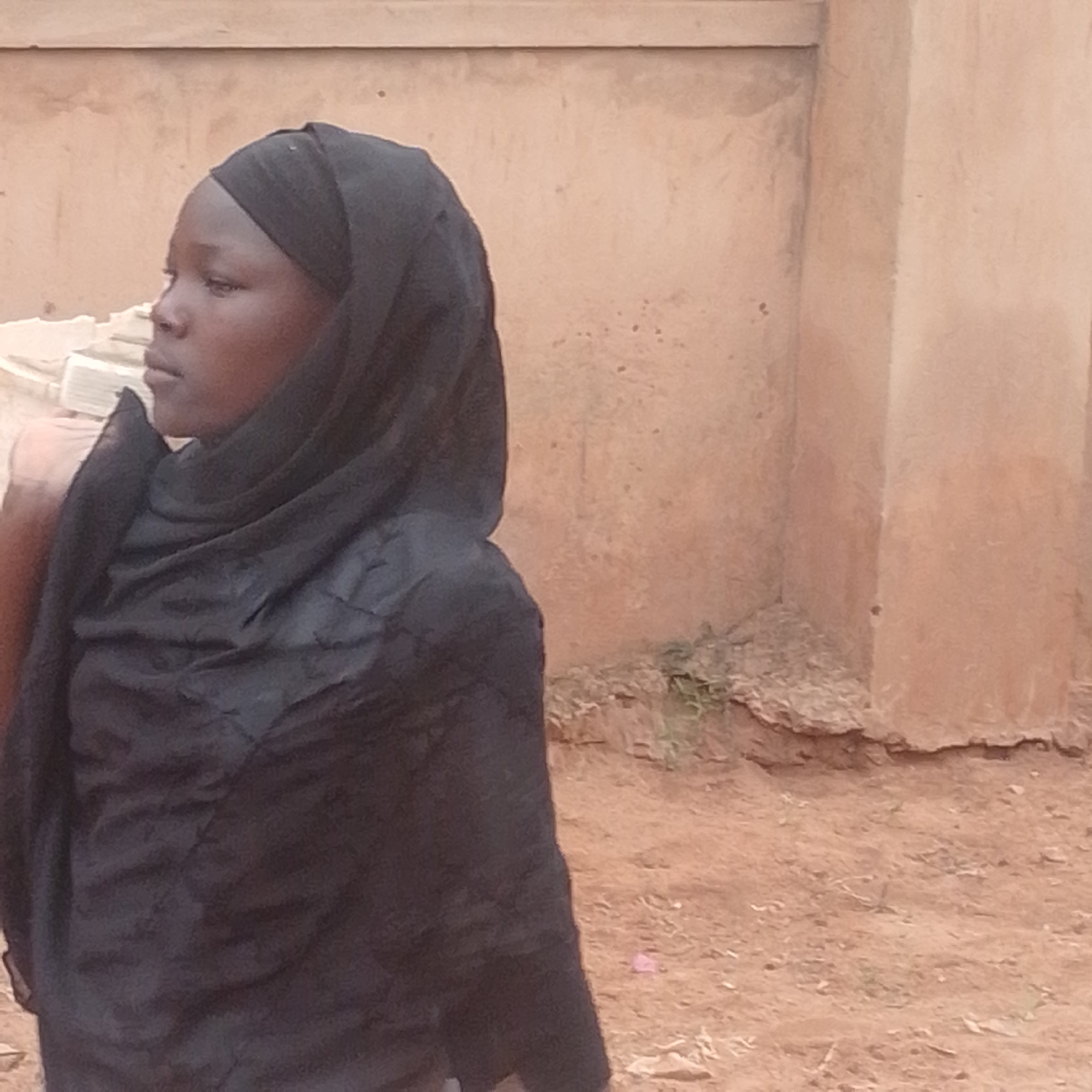 A young girl, wearing a black hijab and dark clothing, is pictured in a profile view, looking to her left. She is positioned in the foreground of the image, with the right side of her face and a portion of her shoulder visible. Her skin tone is dark, and her eyes are closed, suggesting a moment of introspection or rest. The hijab covers her head and neck, with folds of fabric draping over her shoulders.
The background consists of a textured, light brown wall, possibly made of mud or stucco, with subtle variations in color and texture. Below the wall, the ground appears to be a mix of dirt and small stones, with some sparse vegetation growing in patches. The lighting suggests it is daytime, with soft light illuminating the scene. There are no visible objects other than the girl and the natural/built environment. No text is present in the image. The setting appears to be an outdoor environment in a rural or semi-rural area, possibly in Gombe, Nigeria, given the provided location context. The overall mood conveyed by the image is one of quietude and perhaps a touch of melancholy due to the girl's closed eyes and neutral expression.