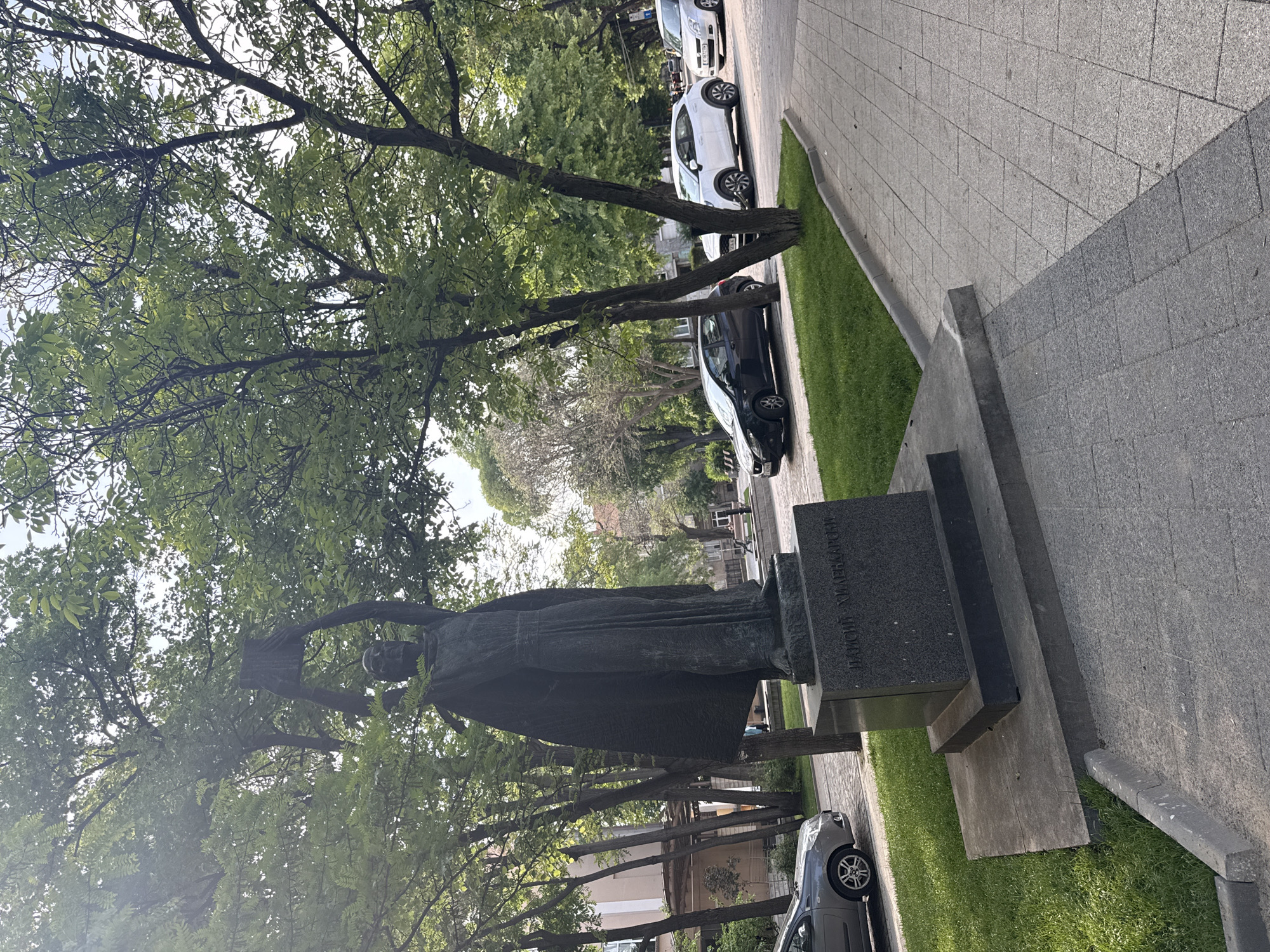 A medium-angle shot depicts a statue in Plovdiv, Bulgaria, obscured by tree foliage. The statue is of a figure in robes holding a scroll and stands on a multi-tiered stone base. "НАСИМ ХИХЛАСОВИ" is inscribed on the base. The statue is situated near a paved sidewalk with a grassy strip bordering it. Cars are parked along the street on the other side of the grass. The sky is visible through gaps in the tree canopy.