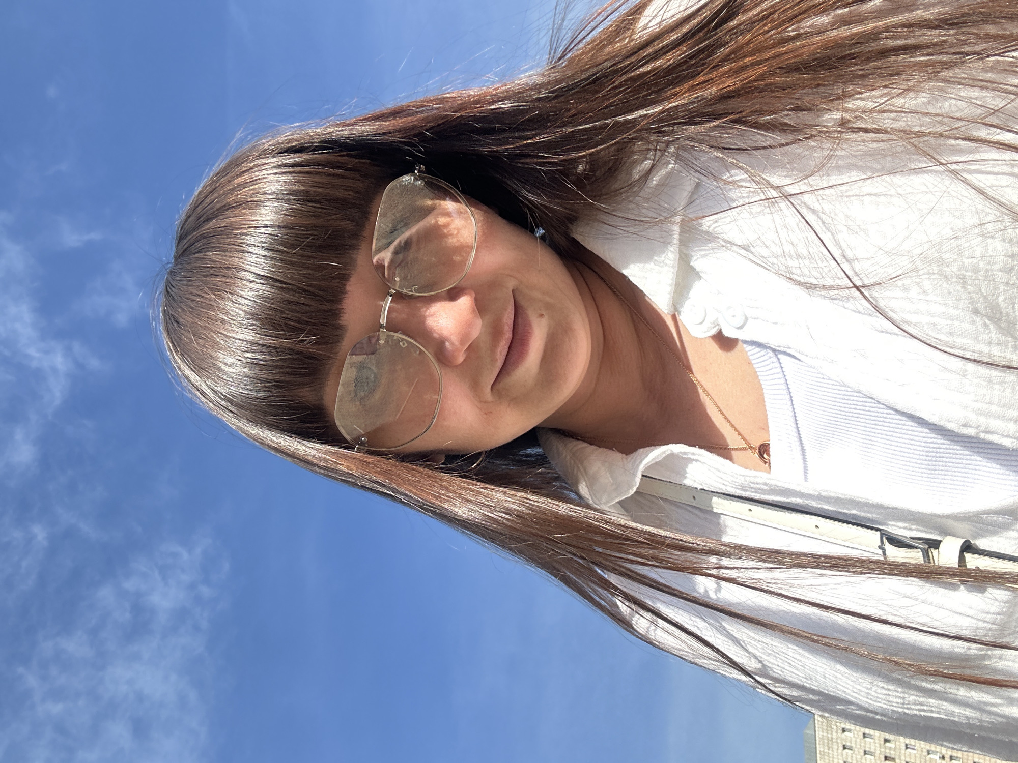 The media file is a selfie taken outdoors, tilted diagonally. The main subject is a young woman with long, dark brown hair, partially obscuring her face. She is wearing glasses with thin, round frames and a white, textured collared shirt, possibly unbuttoned, revealing a lighter blue garment underneath. A delicate gold necklace with a small pendant is visible around her neck. Her hair is windswept, extending across the frame and against a bright blue sky with scattered white clouds. The lighting suggests it is daytime, likely sunny. In the lower right portion of the image, the edge of a building with visible windows and a grey facade can be seen, hinting at an urban or semi-urban setting. The overall mood appears casual and relaxed, possibly capturing a moment of leisure on a clear day. No visible text is present.