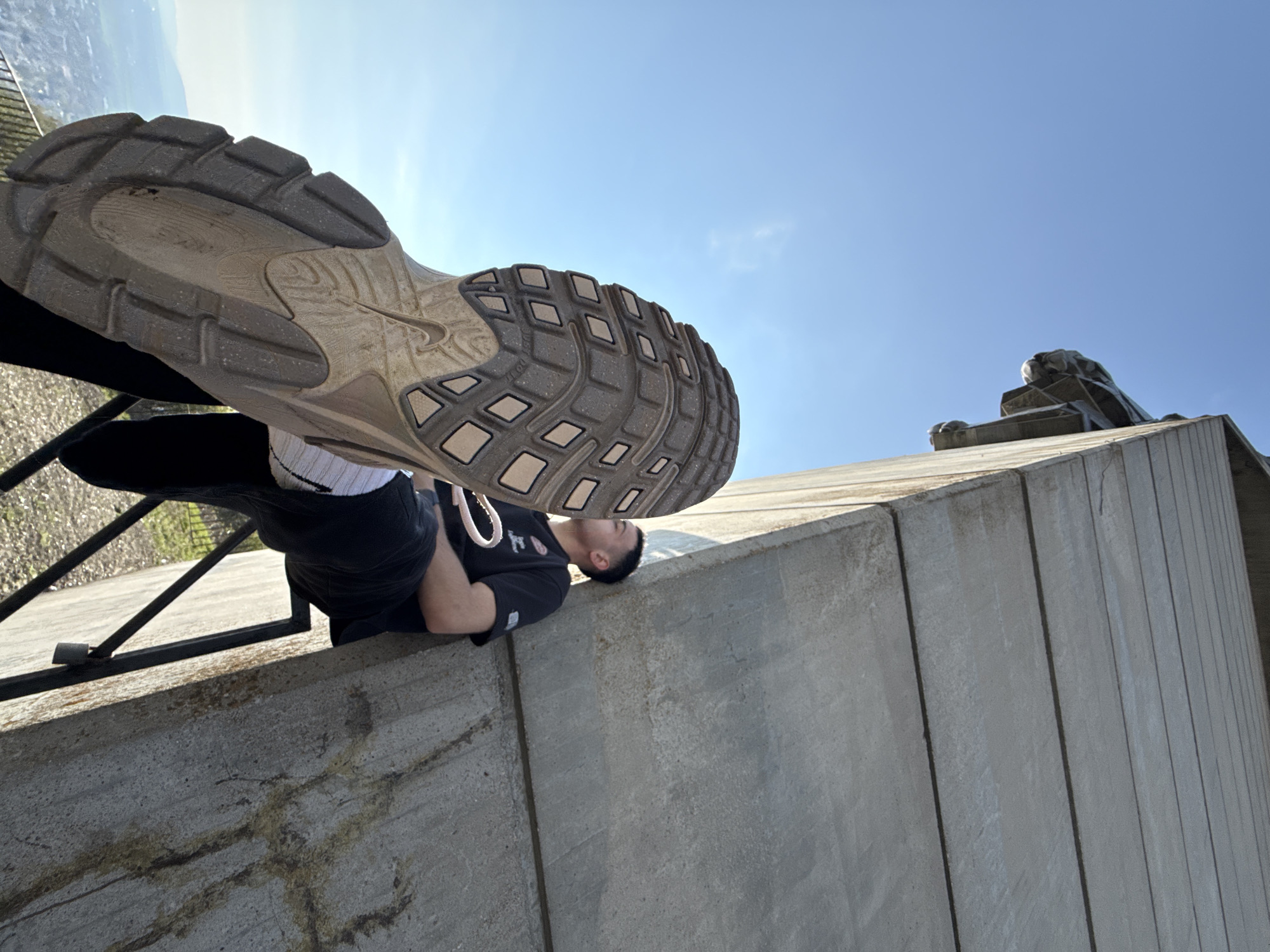 A low-angle, outdoor photograph taken on a sunny day shows a man lying on a concrete ledge with the sole of a shoe prominently displayed in the foreground. The man is lying on his back, wearing a black t-shirt and shorts, and white socks. The shoe, positioned directly above him, is a heavily textured athletic shoe. The concrete ledge is made of large blocks. Behind the man and ledge is a partial view of a railing and an indistinct landscape. The scene appears to be located at the Monument to 1300 Years of Bulgaria in Shumen, Bulgaria, based on the architectural style of the concrete structures and the view of the surrounding terrain.