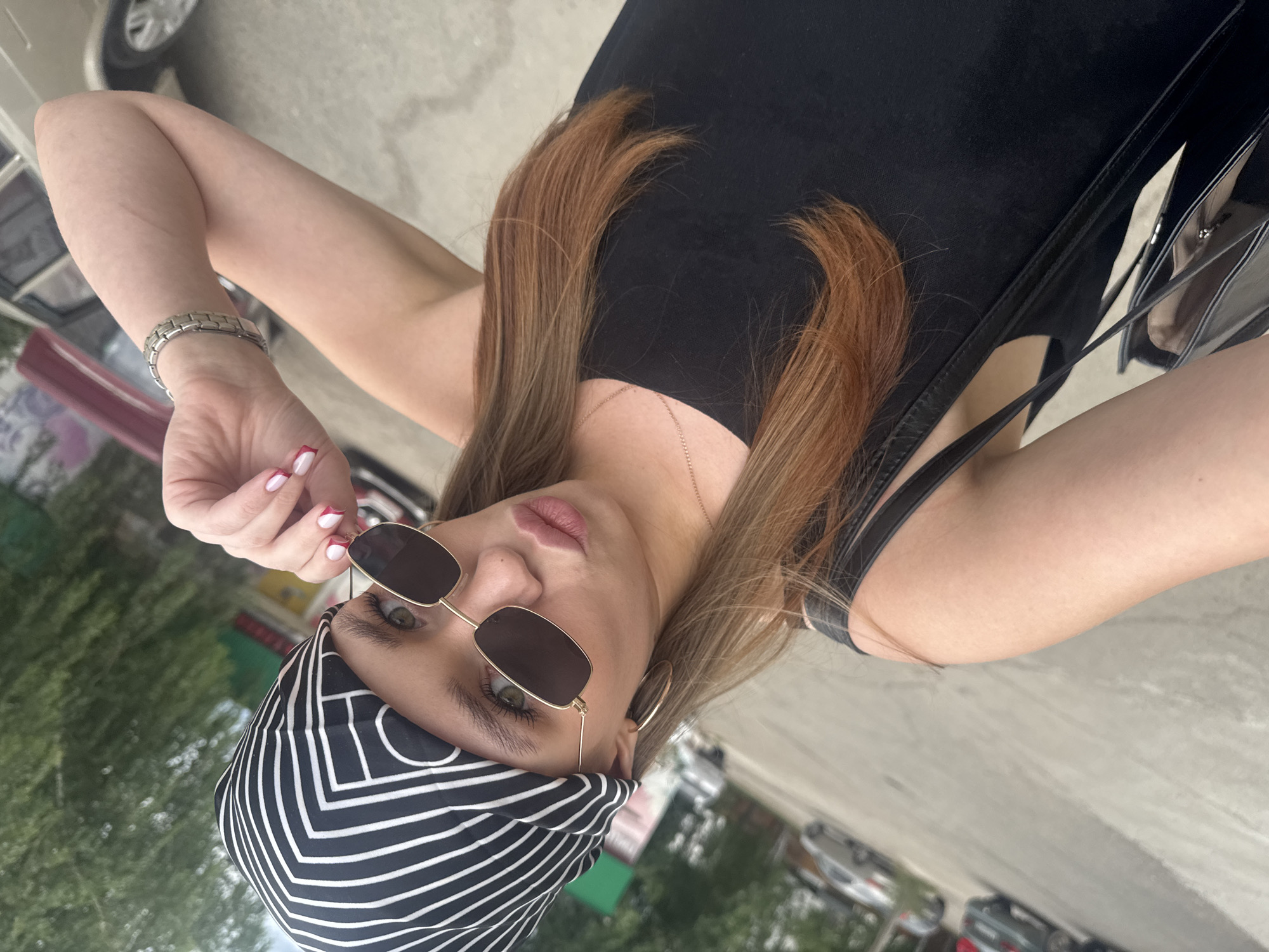 A woman with light brown hair, wearing sunglasses and a black and white striped bandana on her head, is lying down outdoors. She is wearing a black dress and a silver watch on her left wrist. Her right hand is raised and is holding the edge of her sunglasses. The background shows trees, cars, and a paved surface, suggesting an outdoor setting such as a park or street. The lighting indicates it is daytime, and the weather appears to be mild. The woman's expression is neutral as she looks towards the camera. No visible text is present in the image.