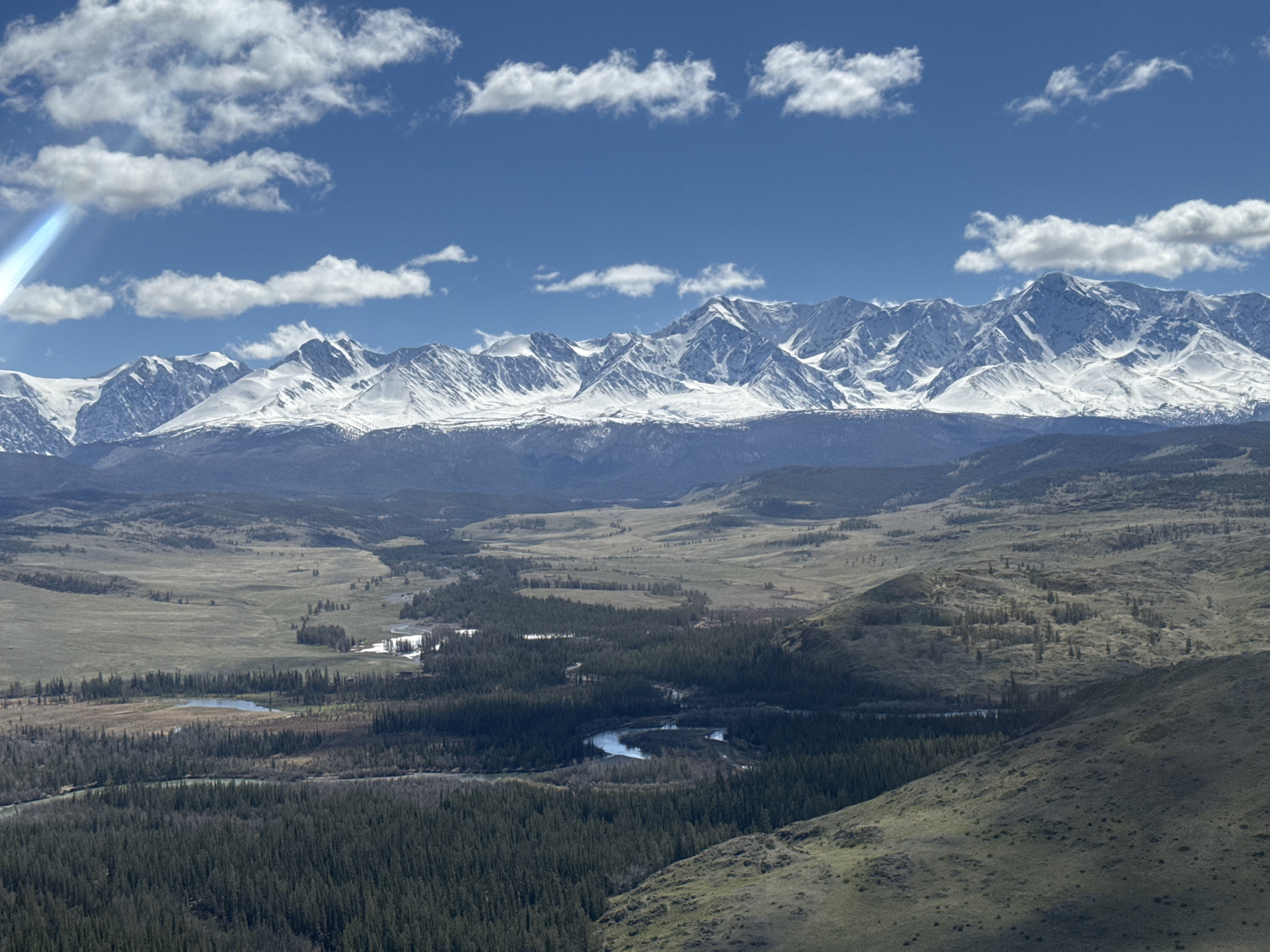 The image shows a mountainous landscape in Kurai, Russia. In the distance, a mountain range with snow-capped peaks is visible under a blue sky with scattered white clouds. Below the mountains, there are forested valleys with a river winding through them. The foreground consists of undulating terrain with vegetation. A lens flare is visible in the upper left corner of the image.
