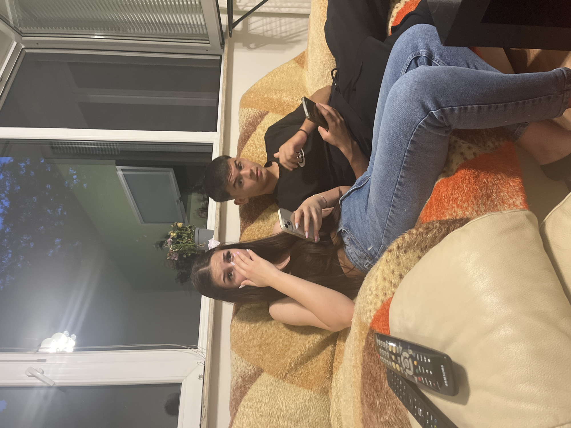 Two individuals, a male and a female, are positioned on a patterned sofa in a room in an unspecified location. The male, wearing a black shirt, is reclining and looking towards the camera while holding a smartphone. The female, dressed in blue jeans and a dark top, also holds a smartphone and appears distressed, covering her face with one hand. The sofa is covered with a textured blanket in shades of brown and orange. In the foreground, two remote controls are visible on a light-colored cushion. A window is present on the left side of the frame, showing an external view of trees and a building. The room is dimly lit.