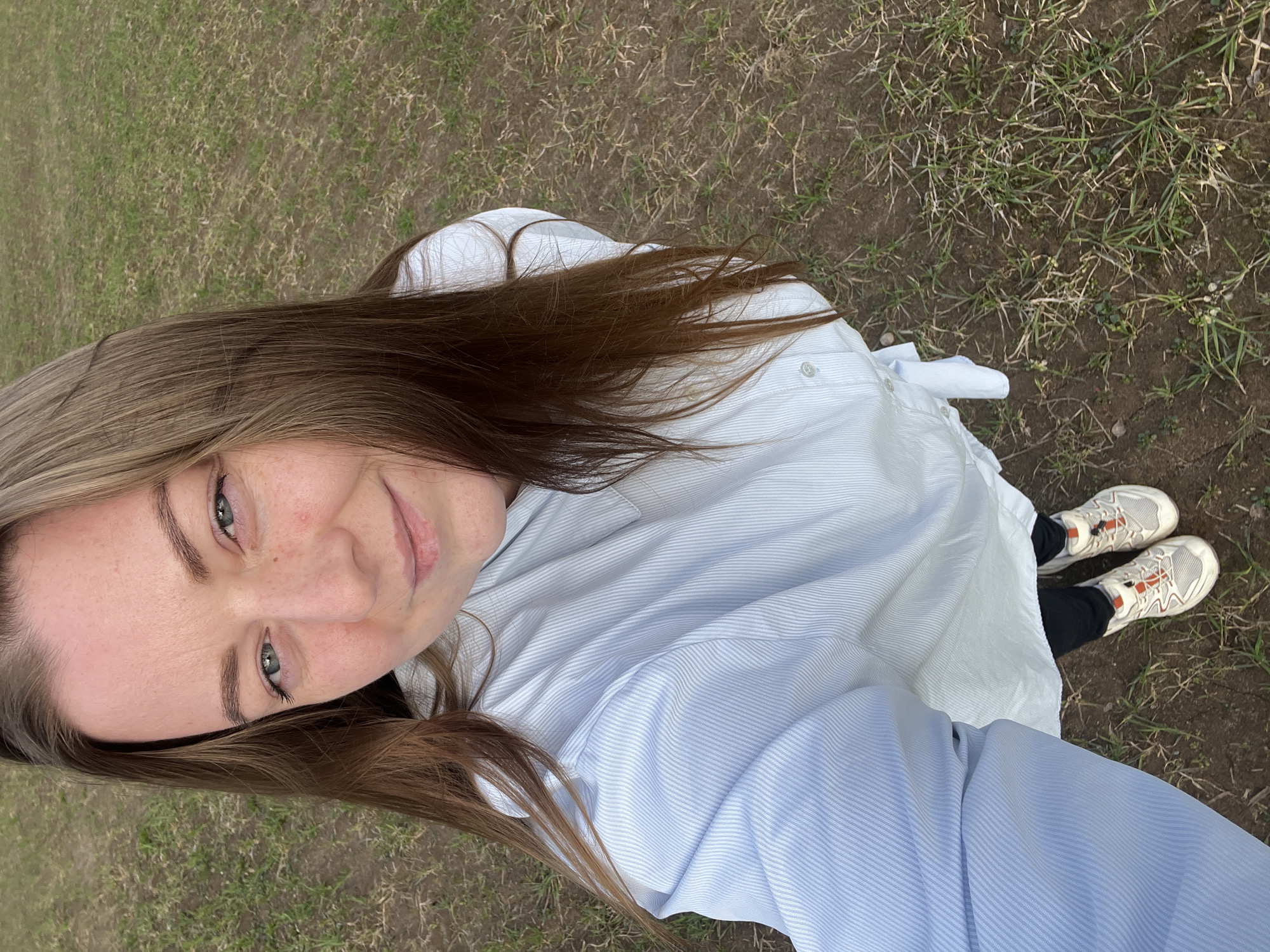 A selfie shot from a high angle shows a woman standing on a patch of grass and dirt. The woman has fair skin, brown hair, and light-colored eyes. She is wearing a light blue striped button-down shirt and black leggings. Her shoes are white and orange athletic sneakers. The ground is covered in sparse grass and bare patches of dirt. The photo appears to be taken in daylight outdoors. The location is presumed to be Раздолье, Russia.