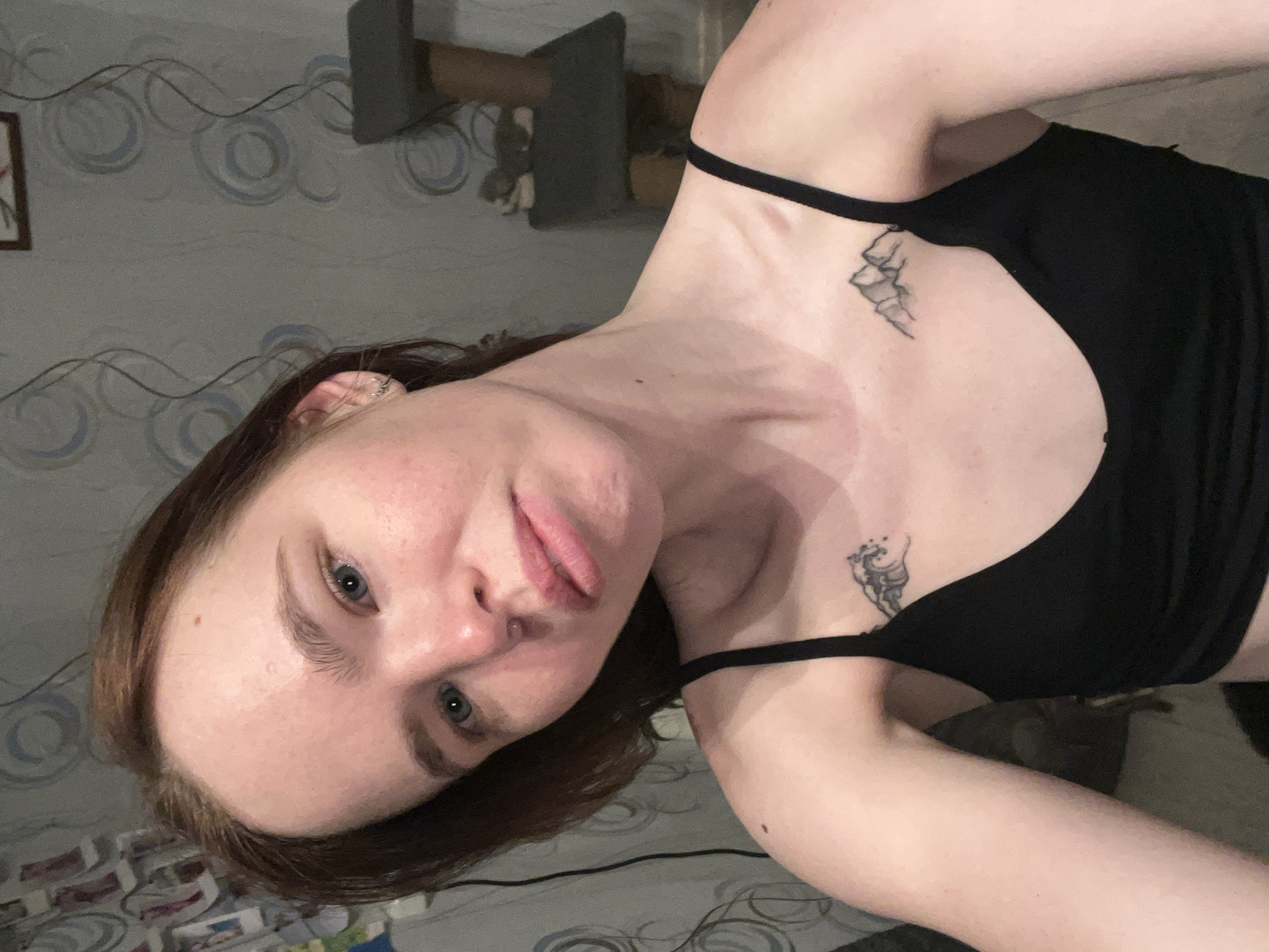 A woman with red hair and blue eyes is centered in the image. She is wearing a black top. She has a tattoo on her right shoulder and another on her chest. The background includes a patterned wallpaper, a cat tree, and other unidentifiable items. The image does not provide information on whether the location is in the city of Chaikovsky, Russia.
