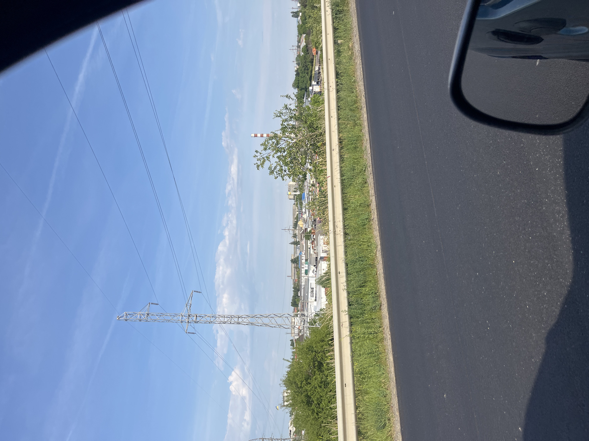 The image shows a roadside view of an industrial area on a sunny day. In the foreground, a black asphalt road is visible, with a white metal guardrail separating it from a grassy embankment. Beyond the guardrail, there are various industrial buildings, some with white roofs and walls, and others appearing to be warehouses or factories. A tall, lattice-like electricity pylon dominates the left side of the frame, with power lines extending across the sky. In the distance, a red and white striped smokestack stands out against the blue sky, which is dotted with white clouds and a faint contrail. Green trees and bushes are interspersed among the buildings. The reflection of a car's side mirror is visible in the lower right corner, indicating the photo was taken from a moving vehicle. The scene appears to be located in an industrial zone on the outskirts of Velyki Lazy, Ukraine, characterized by infrastructure related to power transmission and manufacturing.
