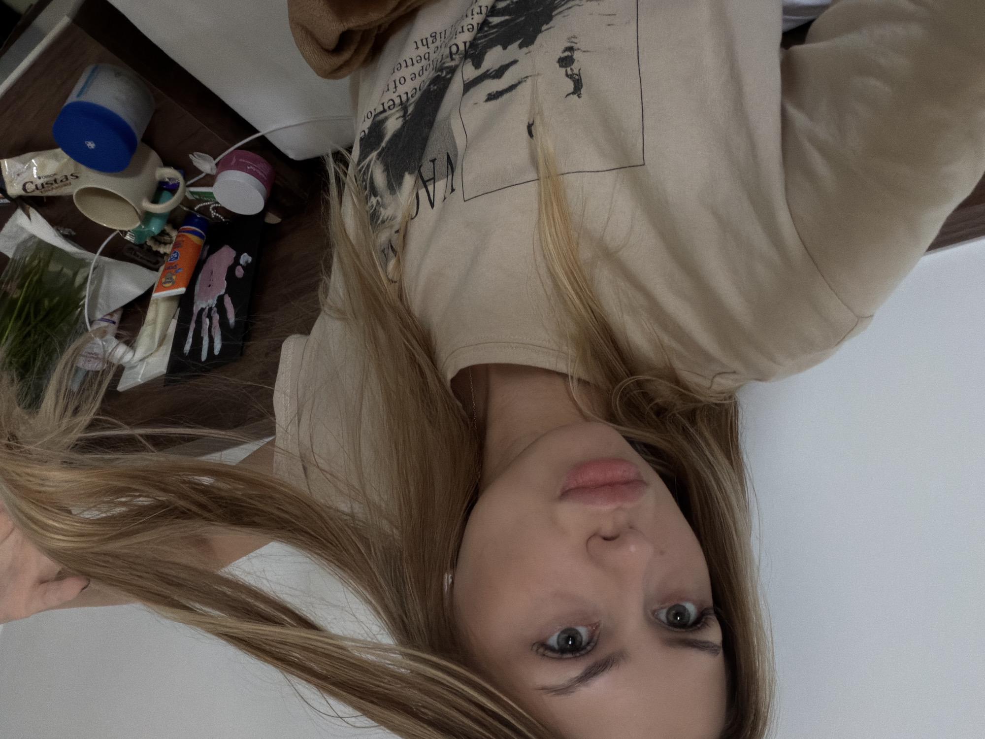 The image shows a young Caucasian woman with long blonde hair, wearing a light-colored t-shirt with a graphic print. She is looking towards the camera with a neutral expression. Her hair is partially obscuring her face. The background appears to be indoors, with a wall on the left and a bedside table or dresser on the right. The bedside table has various items on it, including a mug, jars, a tube of sunscreen, and some other miscellaneous objects. There is a white cable running across the scene. The lighting suggests an indoor setting, possibly a hotel room or bedroom.
The location is not specified in the image itself, and therefore, it cannot be determined to be in the city of Đà Nẵng, Vietnam, or any other specific city.