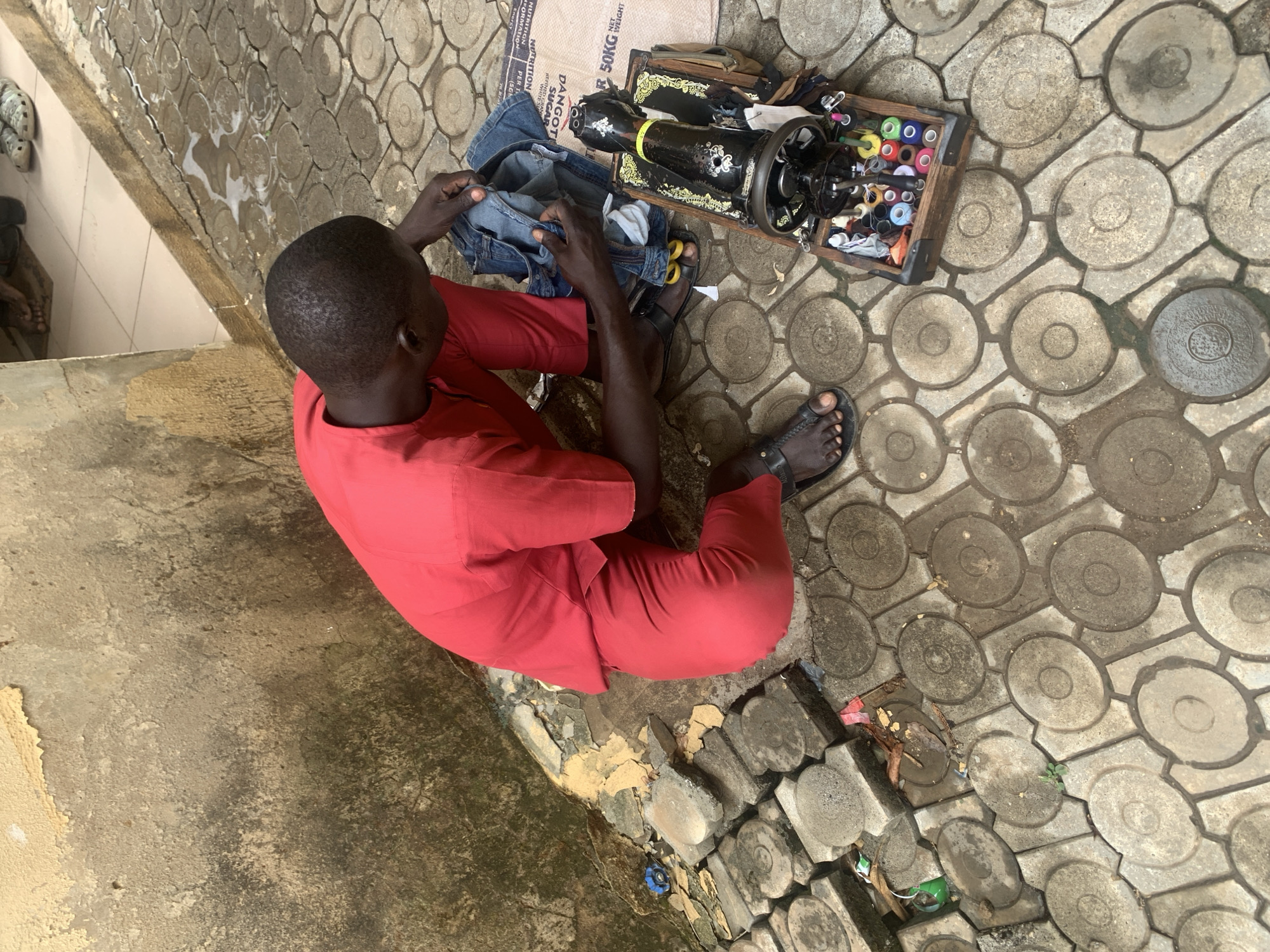 A man sits on the ground outside in Abuja, Nigeria, using a treadle sewing machine. He is wearing a red top and pants and black sandals. The sewing machine is in a wooden box, with thread spools next to it. He holds denim fabric in his hands. A 50kg Dangote Sugar bag sits under the sewing machine. The ground is paved with patterned concrete bricks. The wall near the bottom of the frame has peeling yellow paint.