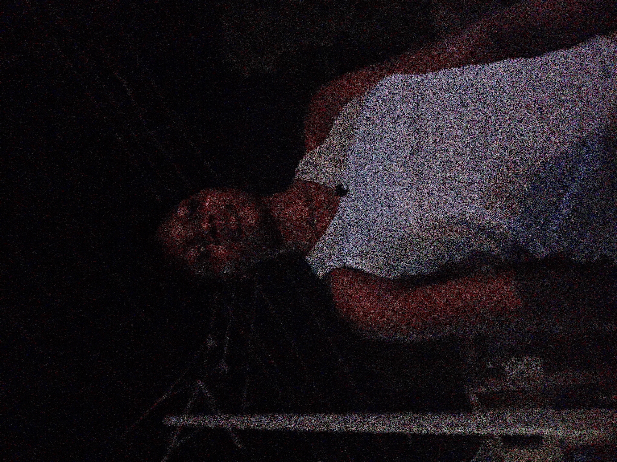 The image is a low-light, grainy photograph of a person wearing a light blue tank top, seen from a low angle, looking upwards. The background is predominantly black, suggesting it was taken at night. To the left of the frame, a vertical pole, possibly a utility pole, is visible, with some overhead wires running across the upper portion of the image. The person's face is partially illuminated, and they appear to be looking directly at the camera or slightly above it. The overall atmosphere is dark and somewhat indistinct due to the image quality and lighting conditions. There are no explicit details to ascertain the exact location within Jalingo, Nigeria, beyond the general context of an outdoor, possibly urban or semi-urban, setting at night.