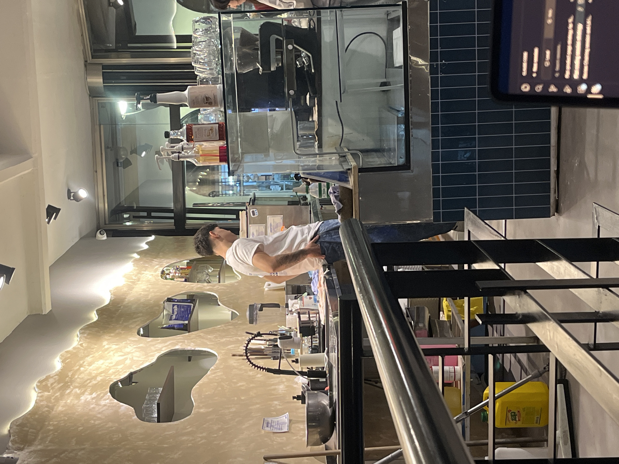 An indoor scene, likely a cafe or bar in Buenos Aires, Argentina, is captured from an elevated perspective, looking down a staircase. The main subject is a man with short dark hair, wearing a white t-shirt and blue jeans, standing behind a bar counter. He faces away from the camera and slightly to the right, with his hands near his waist, appearing to be working or waiting.
The bar area features a modern setup, including a large clear glass enclosure on the counter, showcasing an espresso machine and other equipment. On top of this enclosure are stacked clear glasses, various pump bottles containing colorful syrups (one possibly labeled "MAAN"), and other beverage ingredients. Behind the man, a sink with a flexible faucet and several bar tools are visible.
The wall behind the bar has a unique, textured beige finish with a wavy, organic design, illuminated by integrated lighting along its edges. Irregular-shaped cutouts in this wall reveal darker spaces or rooms beyond. A small white security camera is mounted on the wall.
In the foreground, a black metal staircase with silver handrails descends, partially obstructing the view. Beneath the stairs, a storage area contains several yellow plastic containers, one clearly labeled "CLeZ," likely a cleaning product.
Further in the background, a wall with dark blue tiles is visible. A reflection of what appears to be a smartphone or tablet screen, displaying columns of text or data, is also visible on the right side of the image. The lighting is artificial, suggesting the photo was taken in the evening or at night. The overall atmosphere appears contemporary and functional.