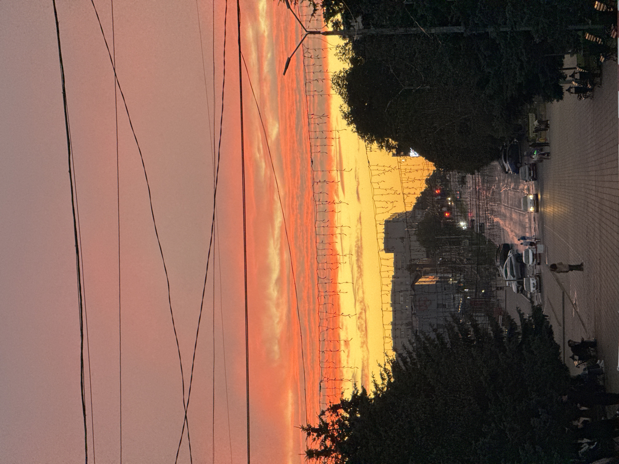The image depicts a street scene in Mikhaylovsk, Russia, during sunset. The sky is filled with vibrant orange and pink hues, indicating the sun is low on the horizon. Overhead, power lines crisscross the sky, and a string of decorative lights hangs across the upper portion of the frame.
The street is paved with cobblestones and slopes downwards into the distance. Cars are parked along the sides of the street and some are in motion. Several individuals are visible on the sidewalks and street, including people walking, standing, and sitting on benches. Trees line the sides of the street, casting shadows on the pavement. Buildings, likely residential or commercial, are visible in the background. The overall atmosphere is calm and serene, characteristic of a quiet evening in a city.