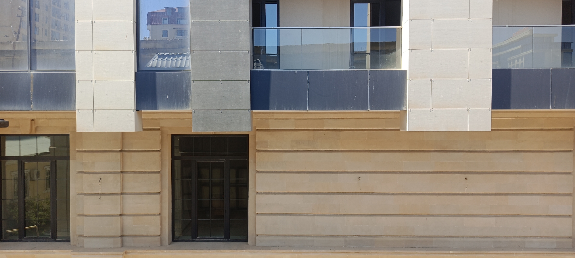 The image depicts the facade of a modern building located in Baku, Azerbaijan. The facade features a combination of materials, including light-colored stone blocks, dark grey panels, and large glass windows. The building has multiple levels, with balconies on the upper floors featuring glass railings. The lower part of the building has a large black-framed glass door. The stone cladding on the lower section is horizontally segmented, creating a textured appearance. The sky above is clear and blue, reflecting in the windows. The overall impression is one of contemporary architecture.