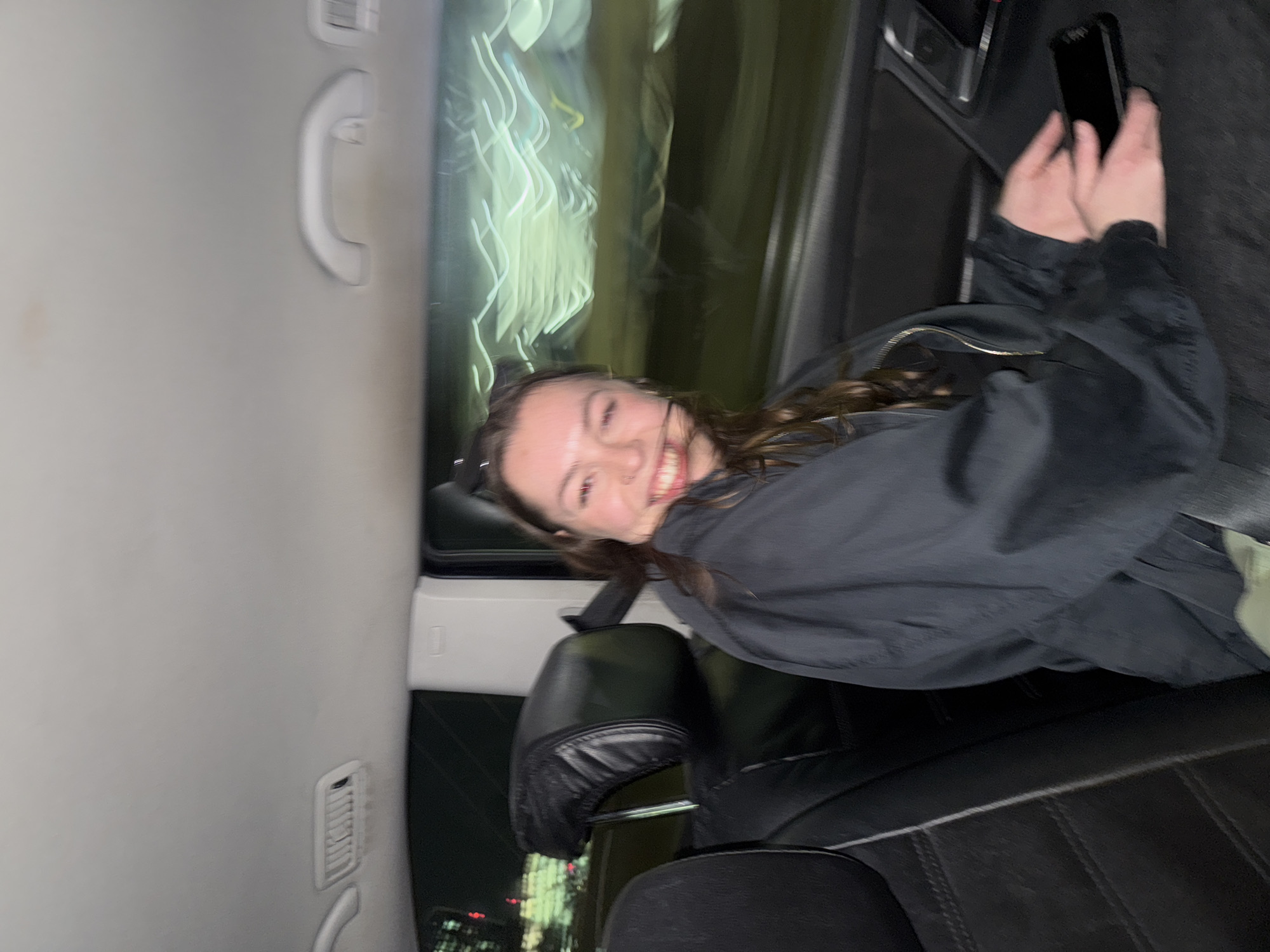 A young woman is captured in a cheerful moment inside a vehicle at night. She is seated, likely in the back seat, smiling broadly with her teeth showing, and looking directly at the camera. Her dark, wavy hair cascades around her shoulders, and she is wearing a dark jacket, possibly black or dark gray. A subtle nose piercing is visible.
Her hands are positioned in front of her, holding a black smartphone horizontally. The vehicle's interior features black leather seats, a light-colored ceiling, and a pillar to her right, which includes a grab handle and an air vent. Through the car window, to her left, blurred streaks of green and white lights are visible, strongly suggesting city lights at night from a moving vehicle, such as in London. The overall lighting is dim, emphasizing her illuminated face against the darker background and the streaky external lights. The image conveys a happy, possibly candid, moment during an evening journey. No discernible text is visible.