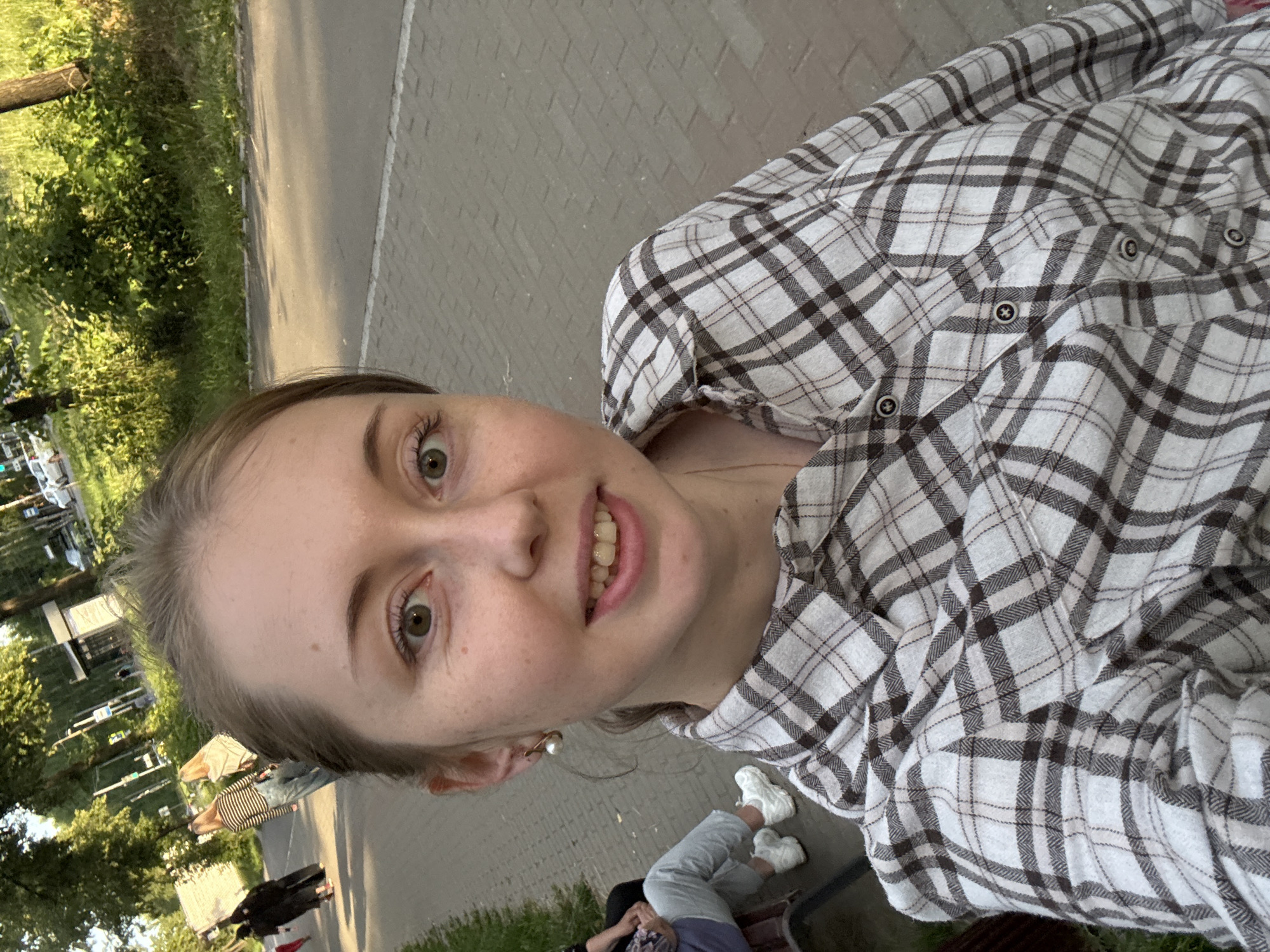 A woman is taking a selfie outdoors. She is wearing a plaid shirt and has light brown hair. Her eyes are open and she is smiling. Her face is in the foreground and takes up a large portion of the frame. In the background, there are trees and people walking. The ground appears to be paved. The image is taken from a high angle, looking down at the subject. It appears to be daytime.