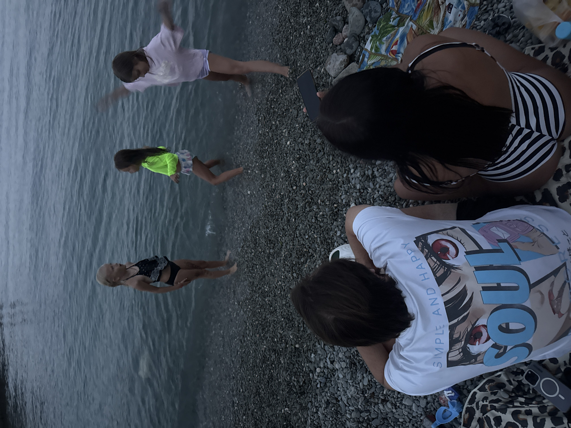 Four people are on a rocky beach next to the sea. The image appears to be taken in the late afternoon or early evening, as the light is soft. The sky is overcast, suggesting a cloudy day with a possibility of rain, but the water is calm. The scene is set in Partenit, Ukraine.
In the foreground, a person wearing a white t-shirt with a large anime-style graphic of eyes and the word "SOUL" is sitting on the pebbles, facing away from the camera. Another person in a black and white striped bikini is also sitting on the pebbles, facing away from the camera.
In the middle ground, three children are in the shallow water. One child in a light purple t-shirt and white shorts is reaching out with both hands. Another child in a bright yellow long-sleeved shirt and a patterned skirt is looking down at the water. A third child, wearing a black and white patterned swimsuit, is floating on their back.
Visible text on the white t-shirt reads "SIMPLE AND HAPPY".