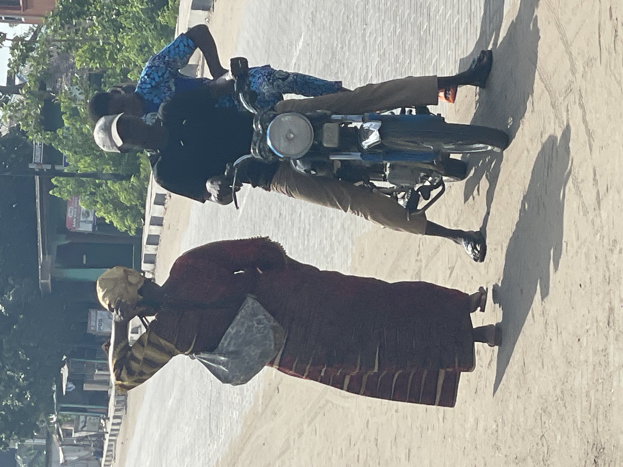 The image depicts an outdoor street scene under bright, clear sunlight, indicated by the strong, distinct shadows cast by the subjects. The setting appears to be an urban or semi-urban environment, with a light-colored, possibly dusty road surface.
In the foreground on the right, two men are on a blue motorcycle that appears to be stationary. The driver wears a dark shirt and a white cap, while the passenger behind him is dressed in a blue patterned shirt. Both are wearing light brown or khaki-colored trousers. The motorcycle is an older model, featuring a prominent circular headlight.
On the left side of the frame, an elderly-looking woman walks. She is wearing a long, dark red or maroon dress with a striped pattern near the hem, a yellow head covering, and carries a grey shoulder bag. Her right hand is raised to her face, possibly shielding her eyes from the sun or adjusting her headscarf.
The background includes green trees and foliage, with glimpses of buildings and utility poles in the distance on the far left. A black and white striped curb runs alongside the road. No readable text is visible. The scene reflects a typical sunny day in a locale consistent with Badagry, Nigeria.