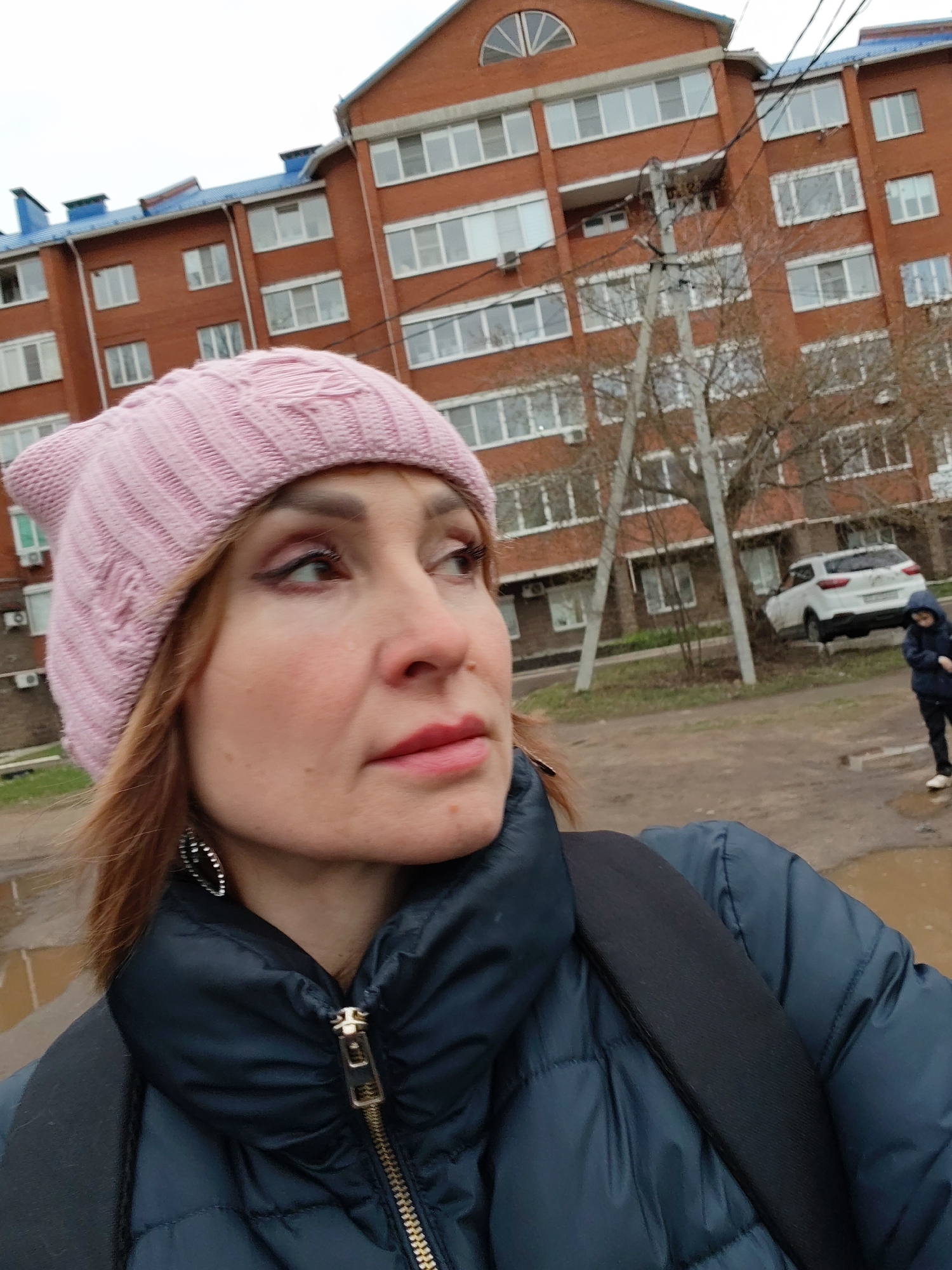 A medium close-up shot captures a woman outdoors, facing slightly to her right and looking away from the camera. She wears a light pink knitted hat with subtle ear-like protrusions, and a dark blue quilted puffer jacket with the zipper partially open, revealing a gold zipper. A black backpack strap is visible over her right shoulder. Her hair is reddish-brown, and she has visible eye makeup, including dark eyeliner and light pink lipstick. She wears a silver, textured oval earring on her left ear.
The background features a multi-story residential building constructed from reddish-brown bricks, with numerous white-framed windows and sections of blue roofing. Several utility poles with electrical wires and bare-branched trees are scattered across the mid-ground. The ground is unpaved, appearing muddy and wet, with visible puddles. A white car is parked in the mid-distance to the right, and a small child in a dark jacket stands near a puddle further back on the right side of the frame. The sky is overcast, suggesting a cloudy day. The scene depicts an urban or suburban environment, consistent with a residential area in Chaikovsky, Russia, during damp weather. No discernible text is clearly visible on the car or elsewhere.